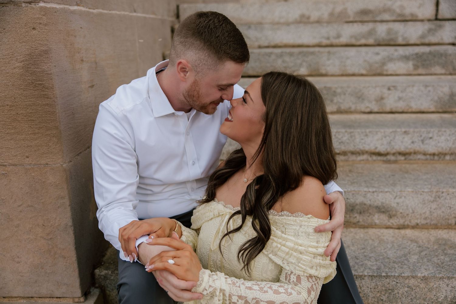 Couple sharing a romantic moment on stone steps, man in white shirt embracing woman in lace off-shoulder top.