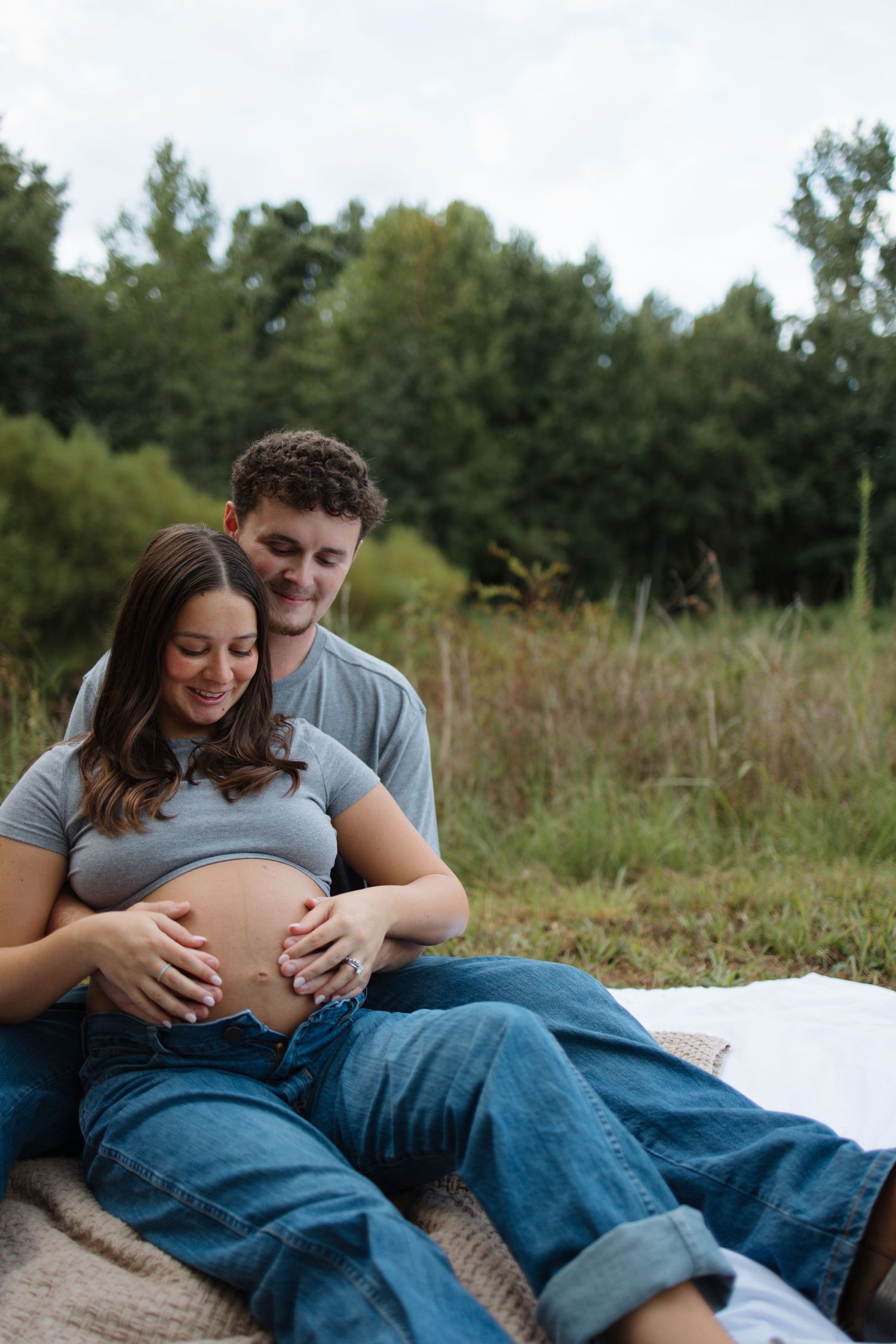 A couple relaxes on a white blanket in a grassy field during a maternity photoshoot.
