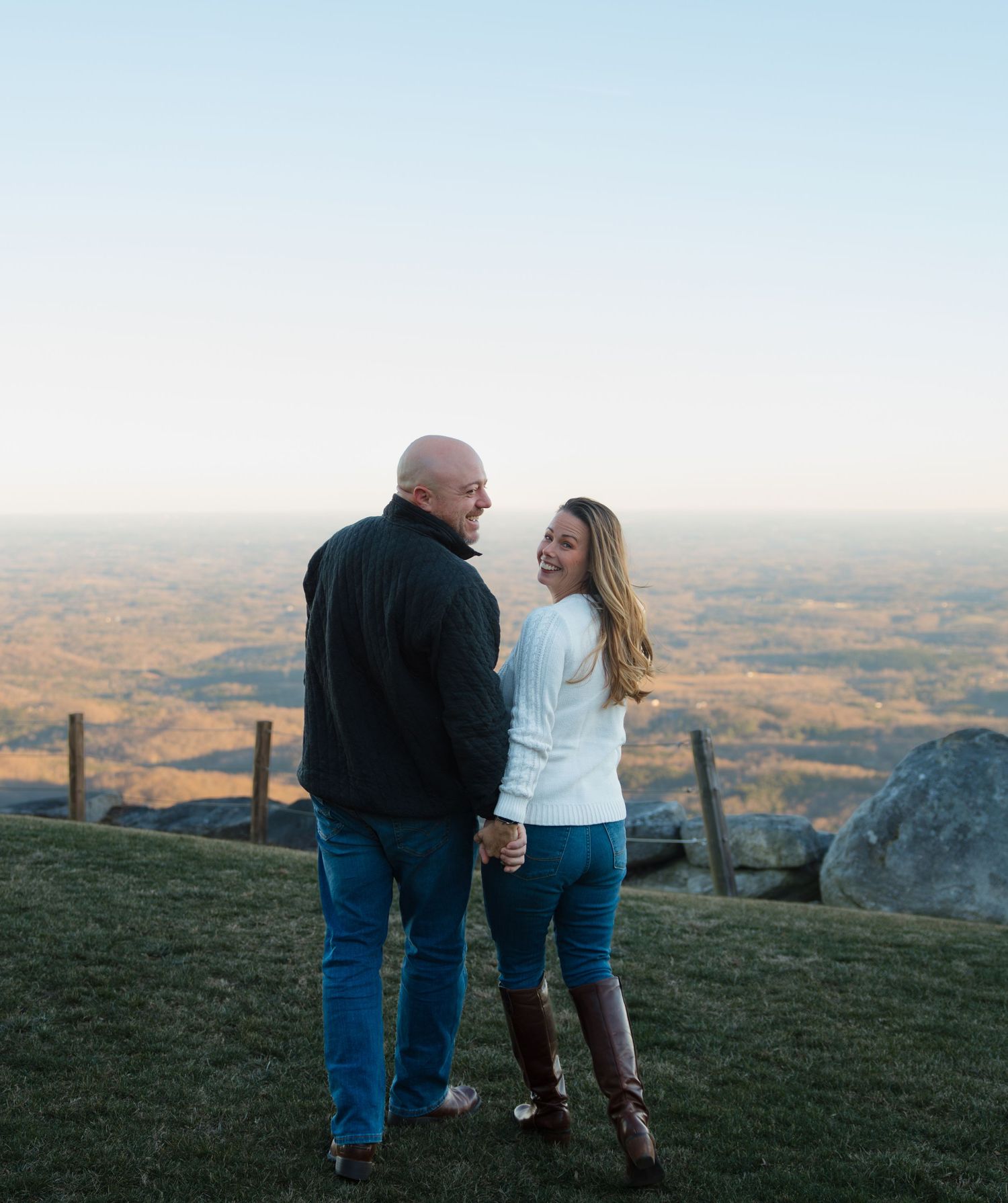 A couple stands close together on a hilltop overlooking a vast autumn landscape.