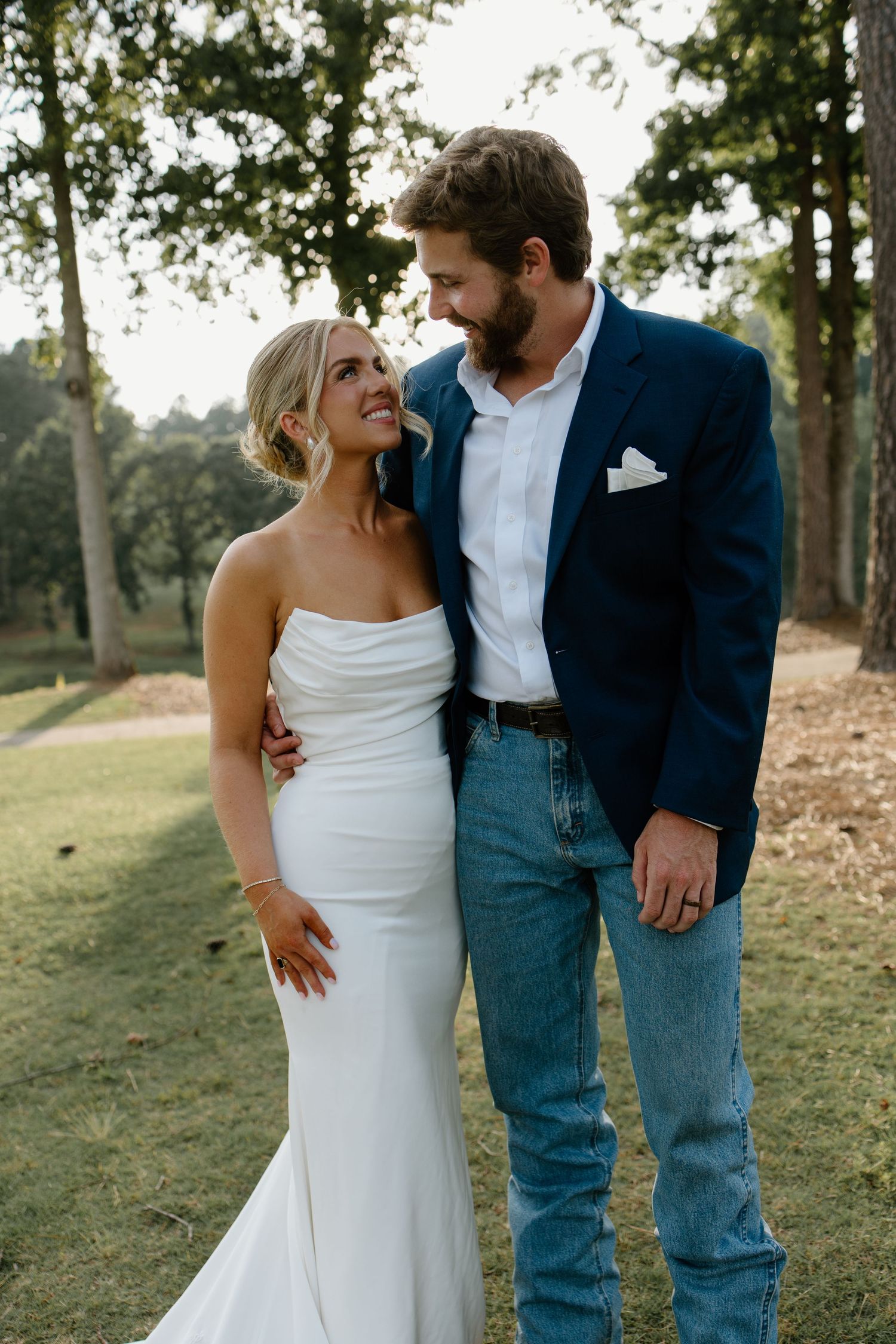 A couple shares a romantic moment outdoors, with the bride in a fitted white gown and groom in navy blazer and jeans.