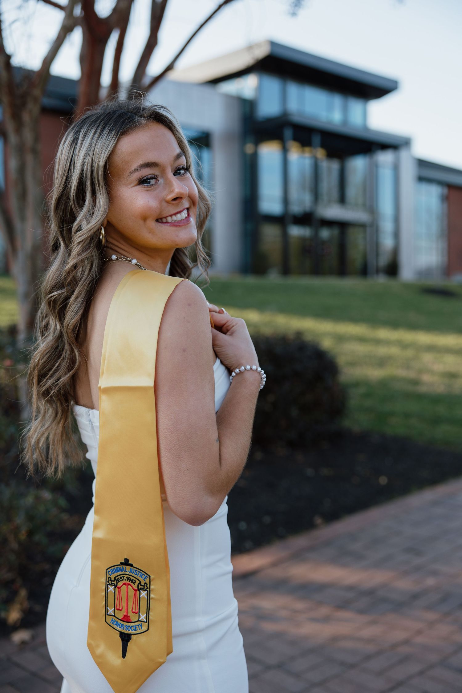 Graduate wearing white dress and gold sash poses with graduation stole outdoors.