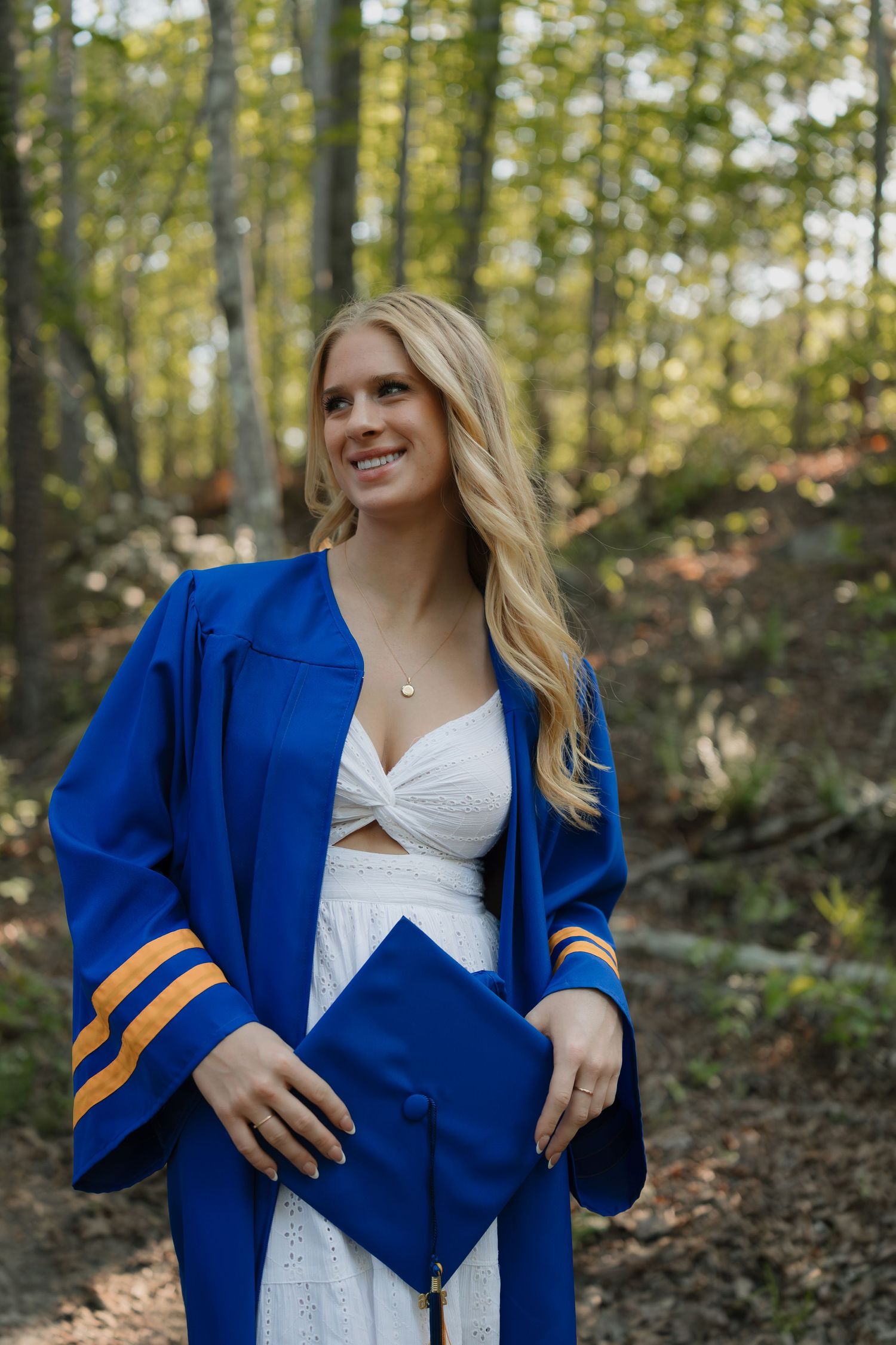 Smiling blonde graduate in blue and gold cap and gown poses outdoors in a forest setting.