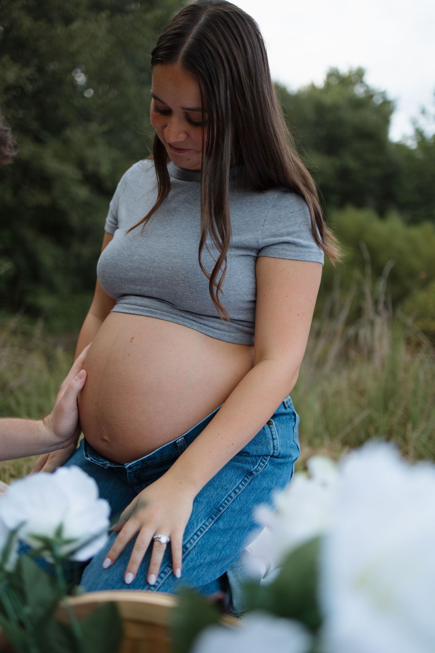 Maternity portrait showing pregnant woman in jeans and grey top with white flowers.