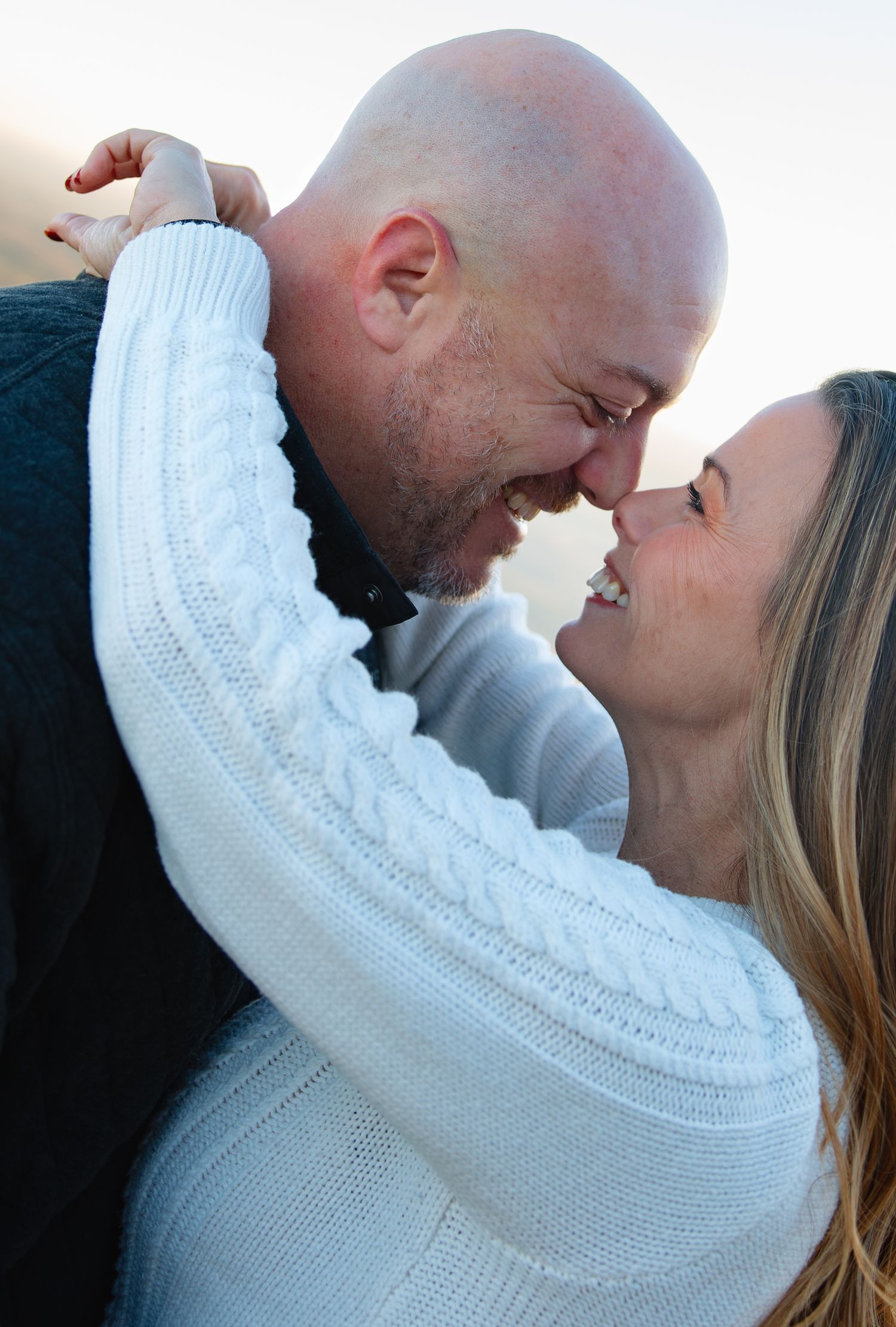 A couple in warm sweaters shares an intimate moment against a sunset sky.