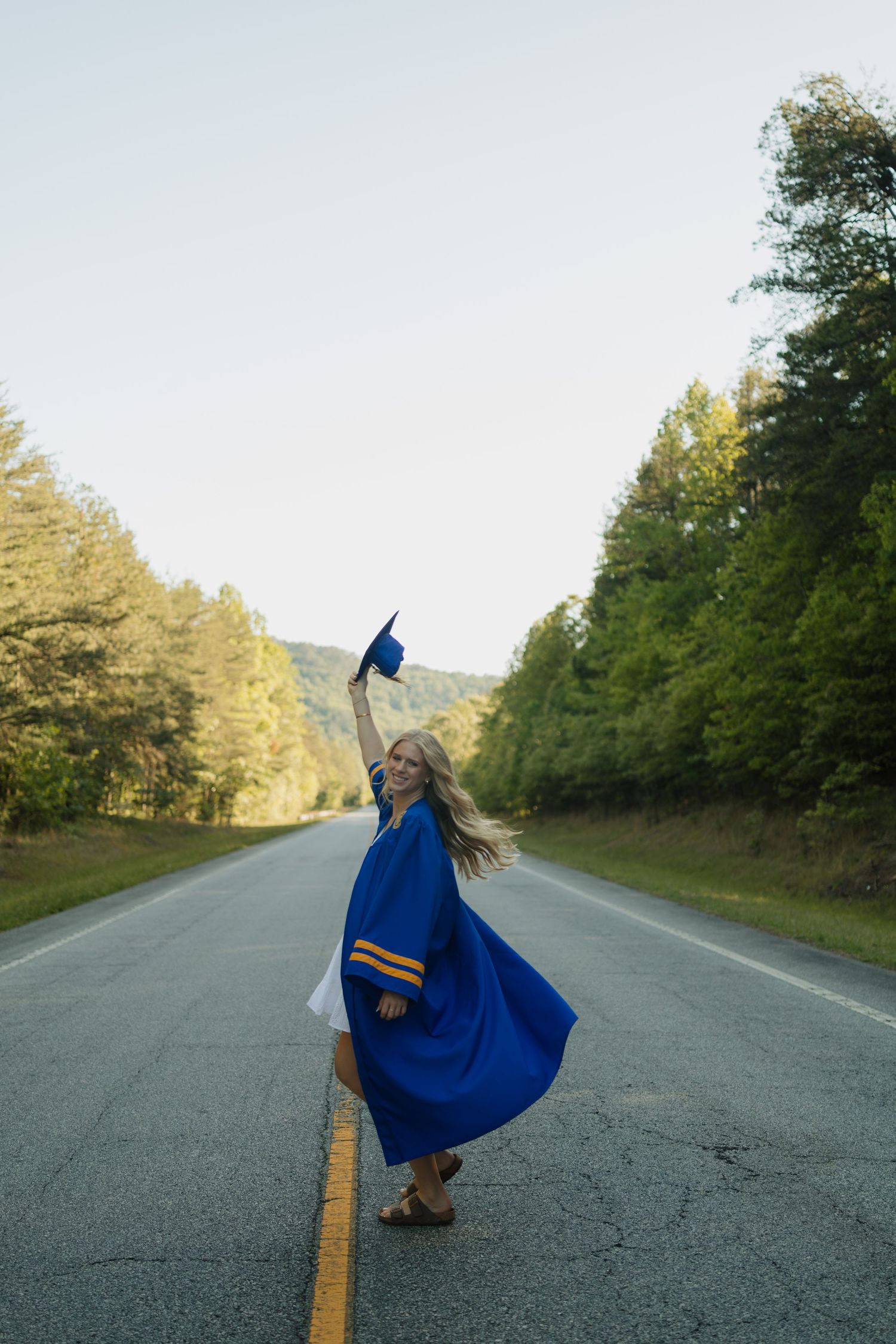 Graduate in blue cap and gown celebrating on a scenic tree-lined road, tossing her cap in the air.