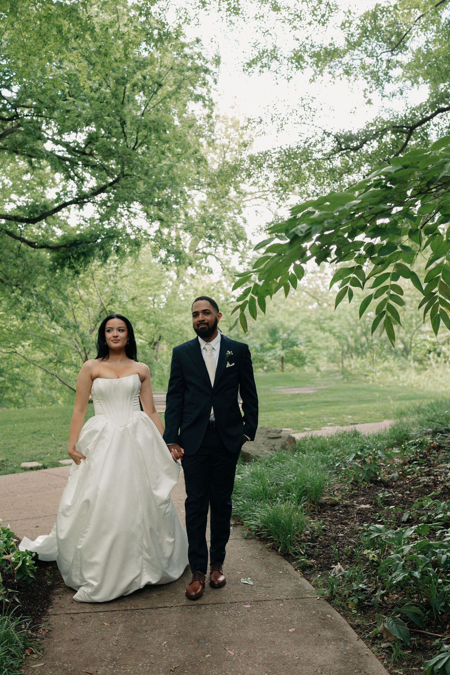 A bride in a white ballgown and groom in a dark suit walk hand in hand along a garden path surrounded by lush green trees.