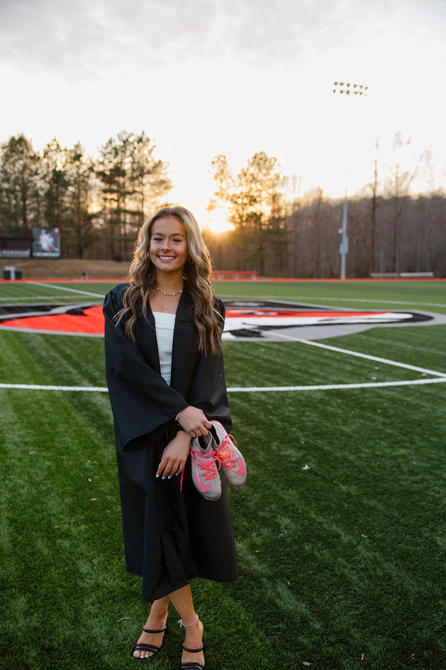 Graduate in black cap and gown poses on sports field at sunset holding pink shoes.