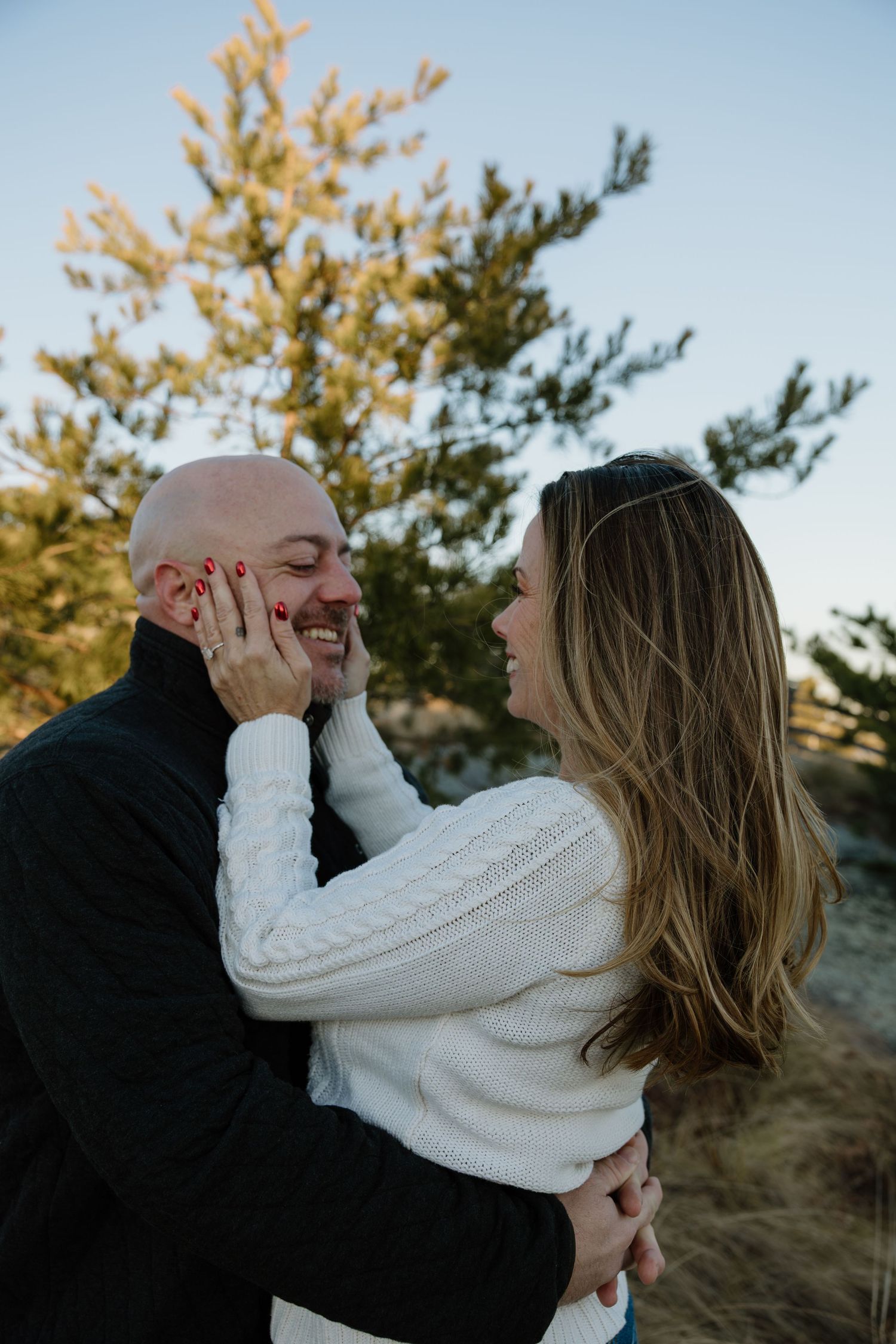 A couple shares an intimate moment outdoors wearing cozy sweaters against an autumn backdrop with golden trees.