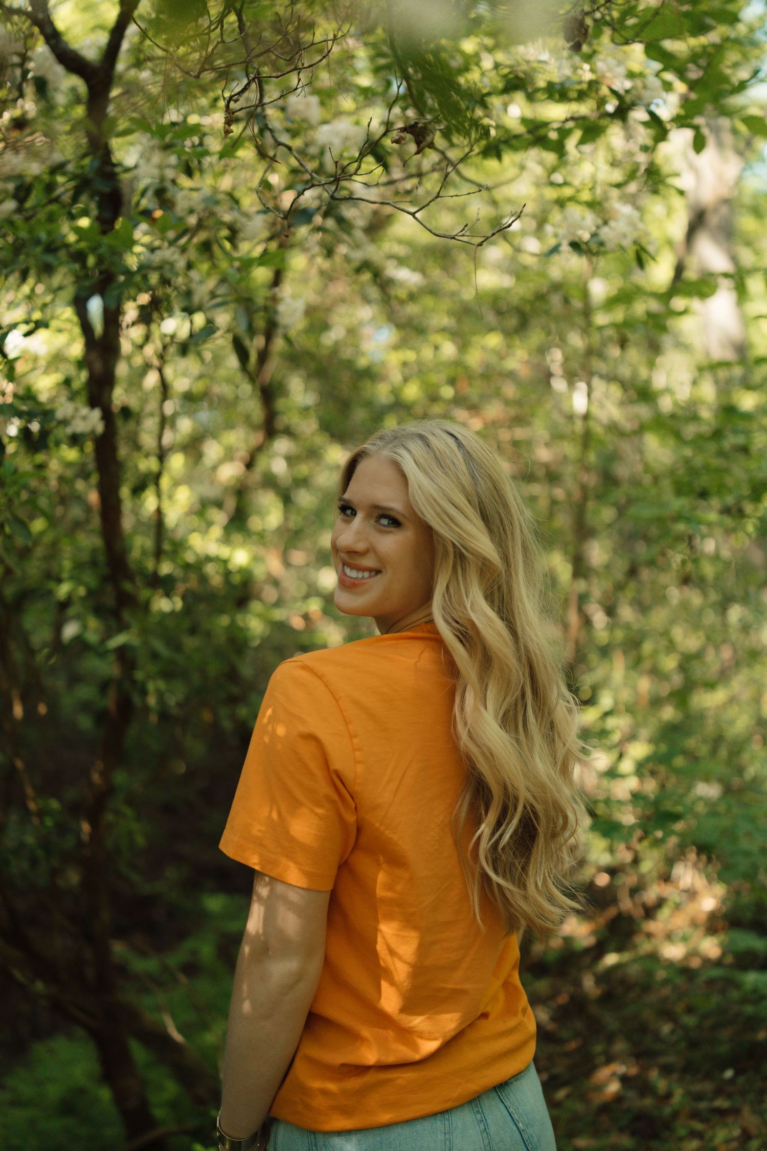 Smiling blonde woman in orange t-shirt turning back in lush green forest setting.