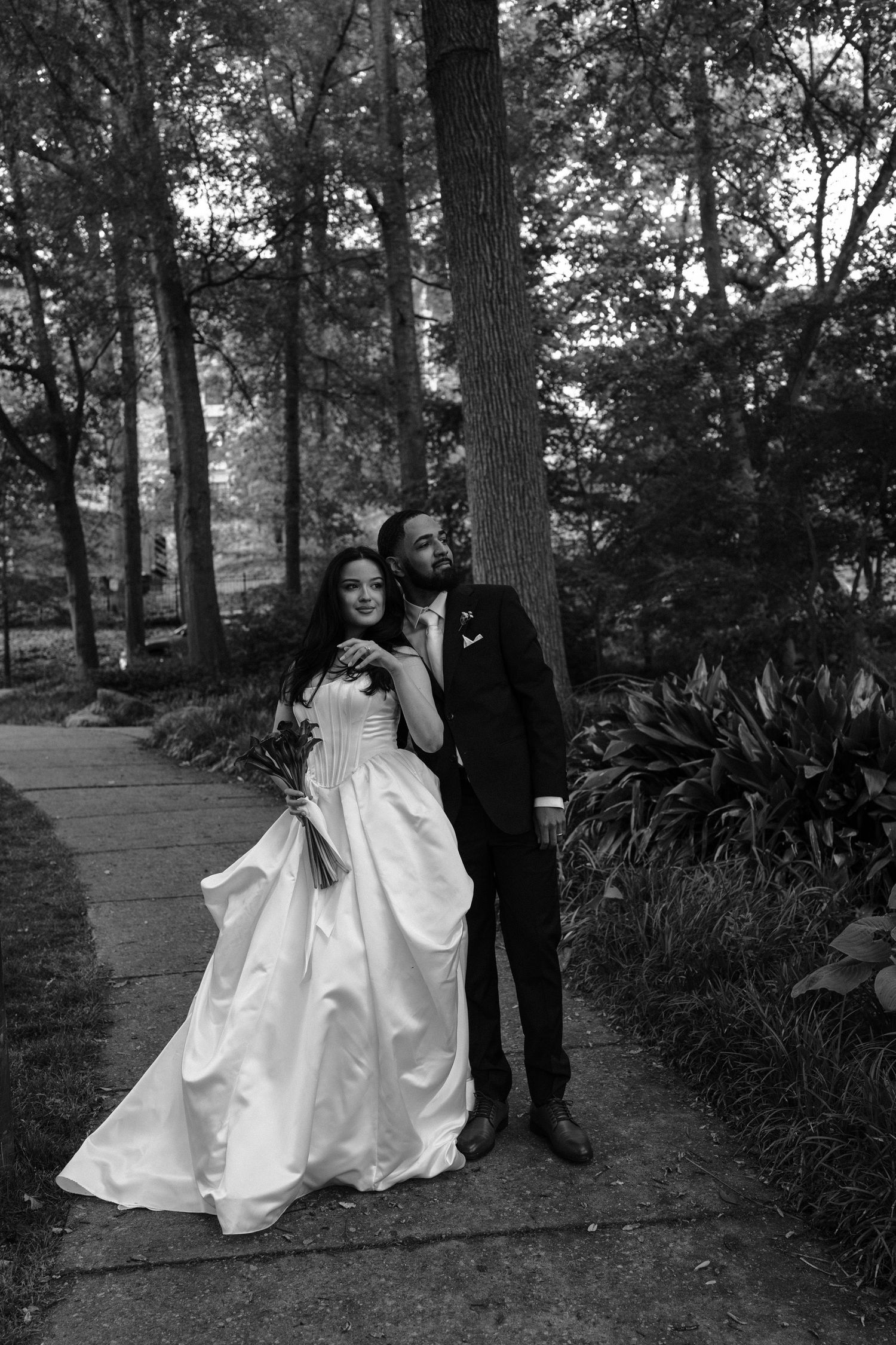 Black and white photo of a bride and groom embracing on a garden path surrounded by tall trees.