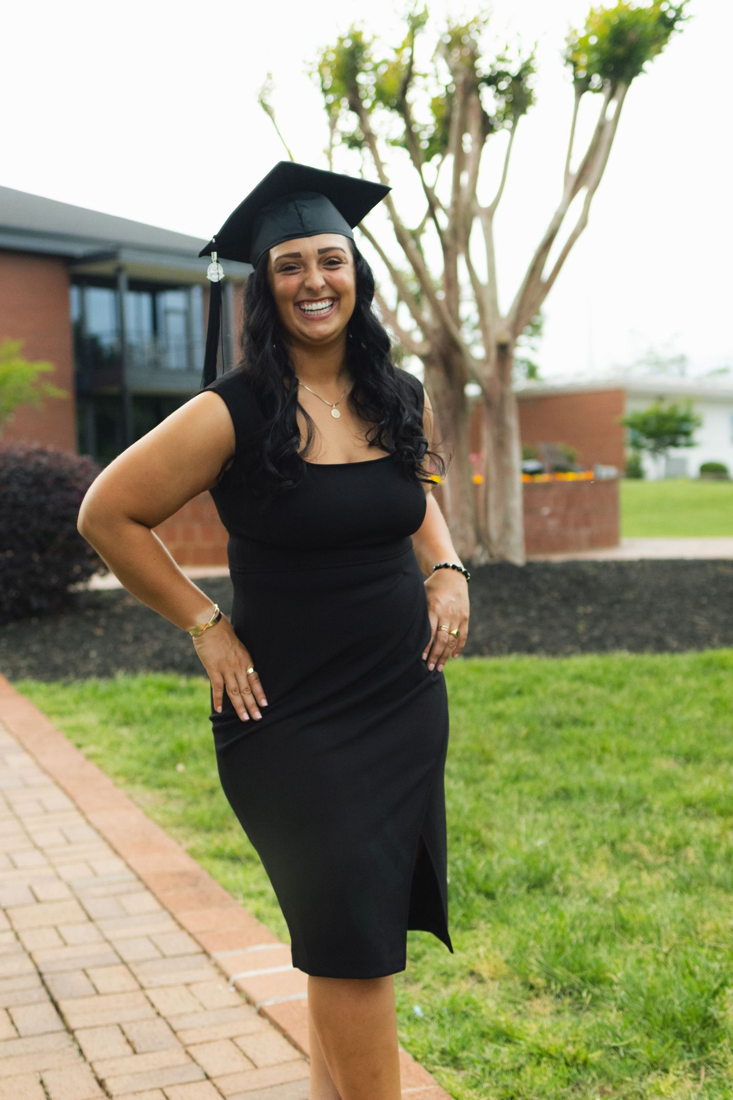 Graduate in black dress posing outdoors on brick pathway with modern building in background.
