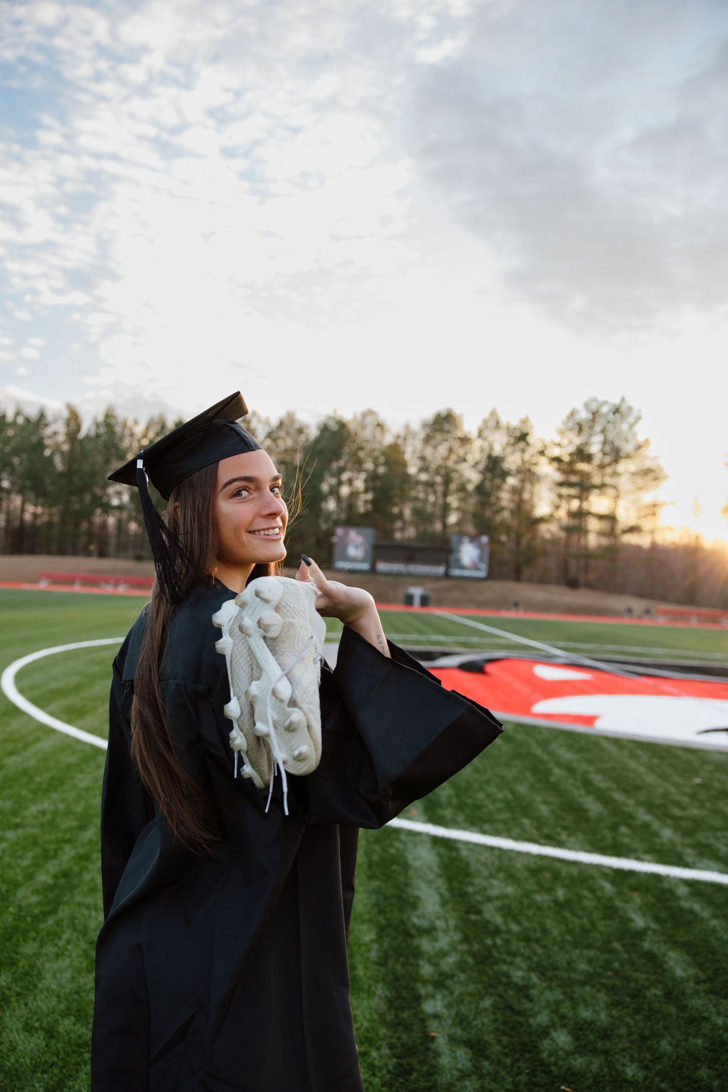 Graduate in cap and gown celebrates on sports field during golden hour.