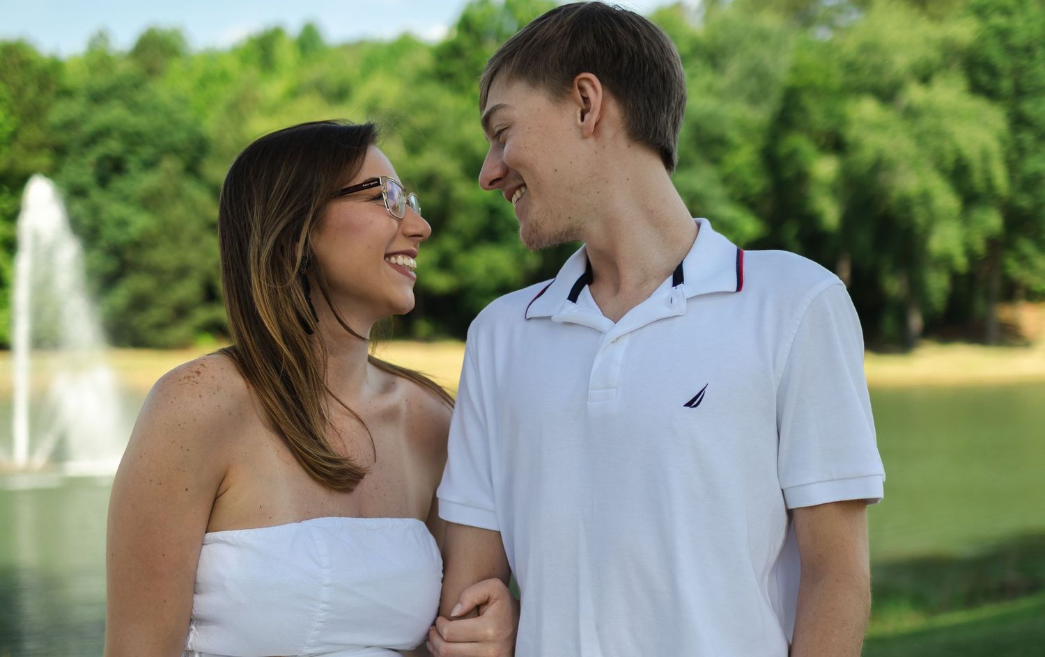 Couple in white outfits laughing and embracing while walking together in a lush green park setting.