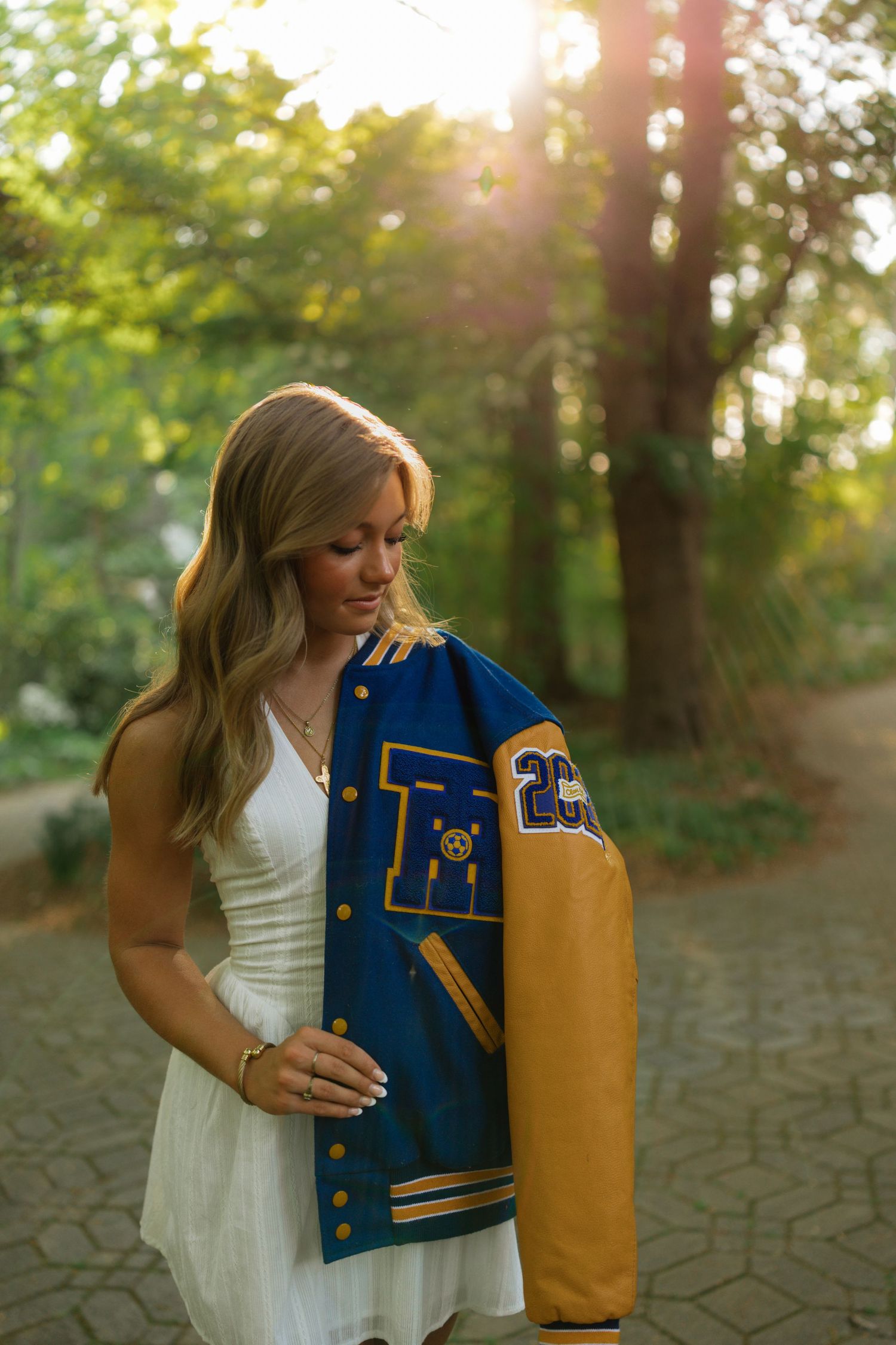 Young woman in white dress holding a blue and gold varsity letterman jacket outdoors in golden hour sunlight.