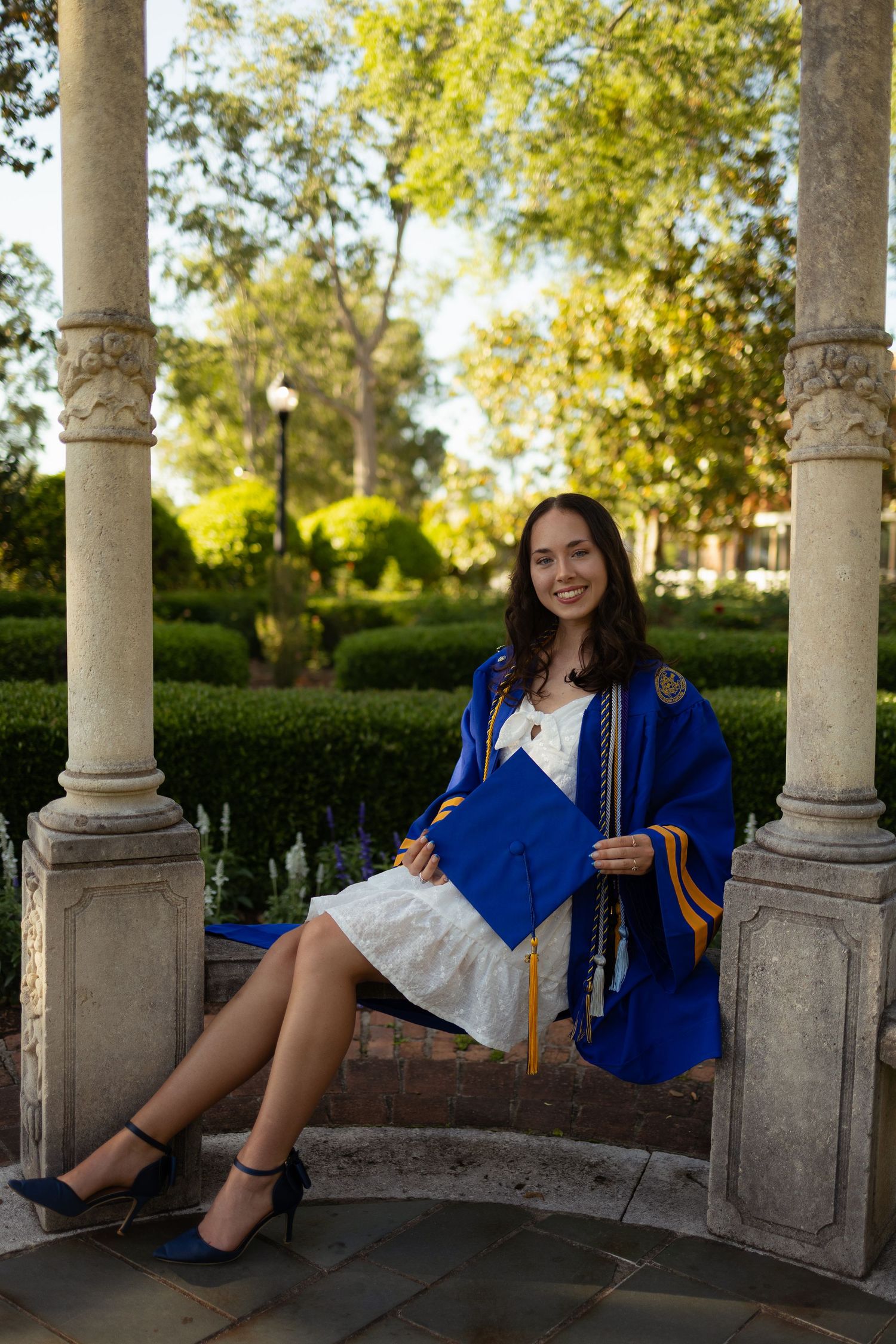 Graduate in royal blue cap and gown poses in outdoor colonnade with stone pillars during sunny graduation photoshoot.