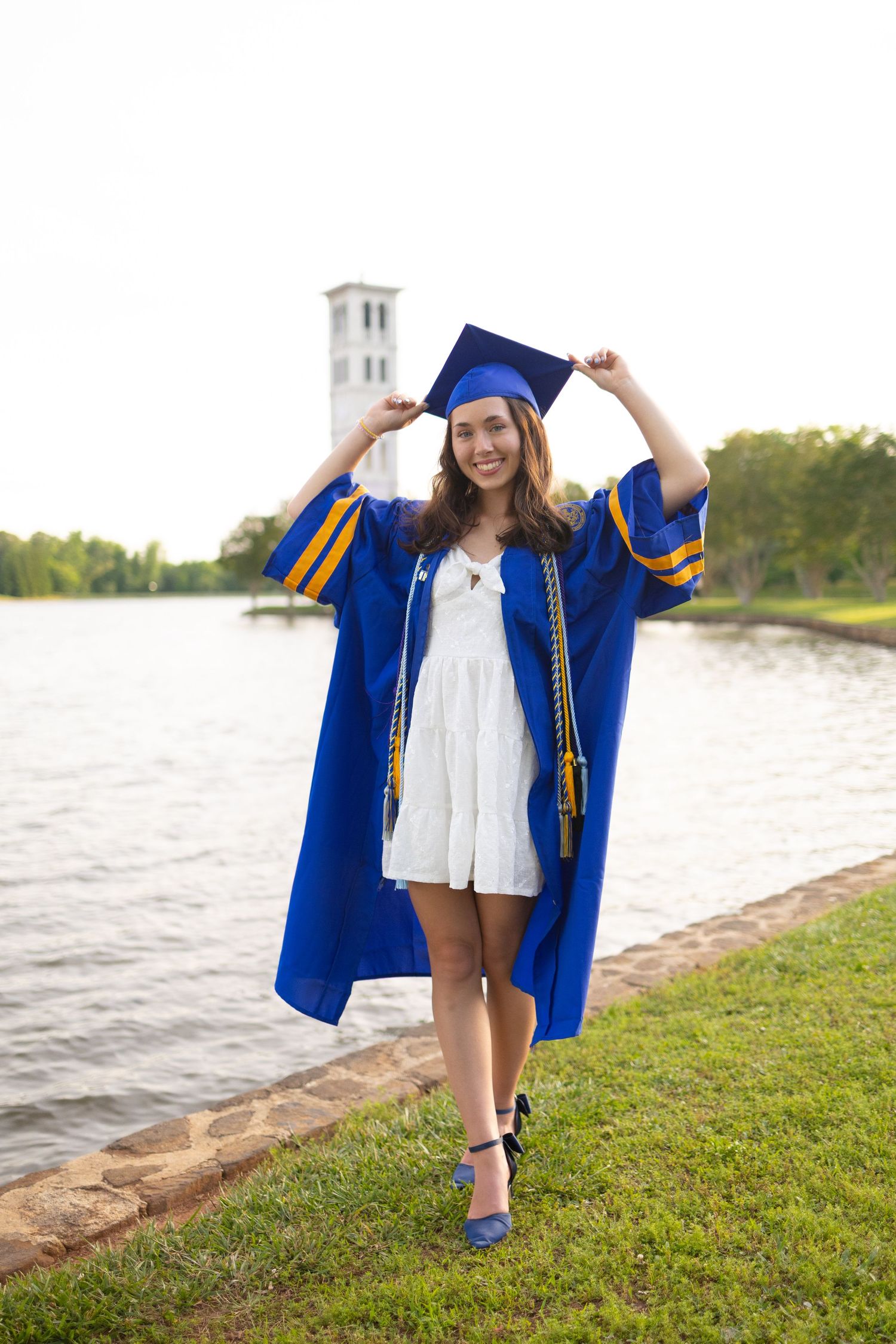 Graduate in blue cap and gown twirling by waterfront with church spires in background.