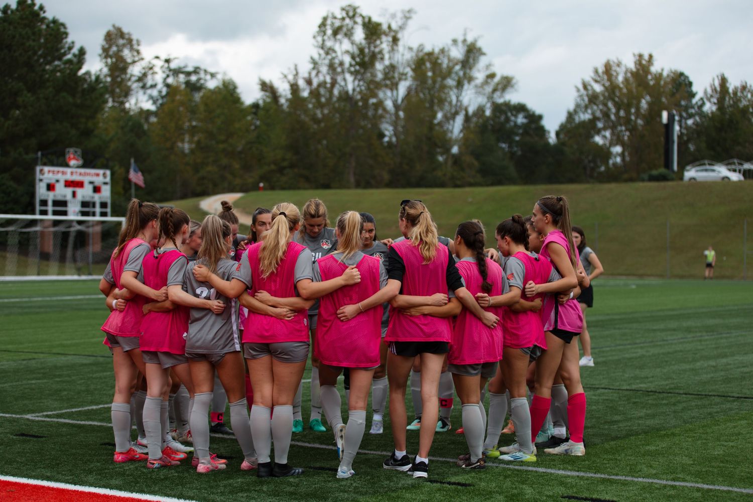 Soccer team forms unity circle wearing pink uniforms on sports field.