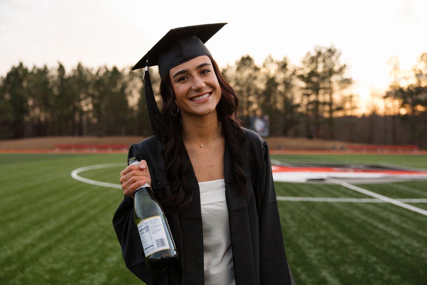 Graduate in black cap and gown celebrates on sports field at sunset.