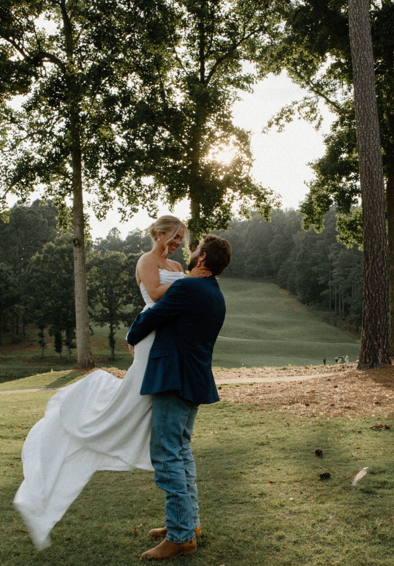 A romantic couple dancing together on a grassy lawn at sunset with trees in the background.