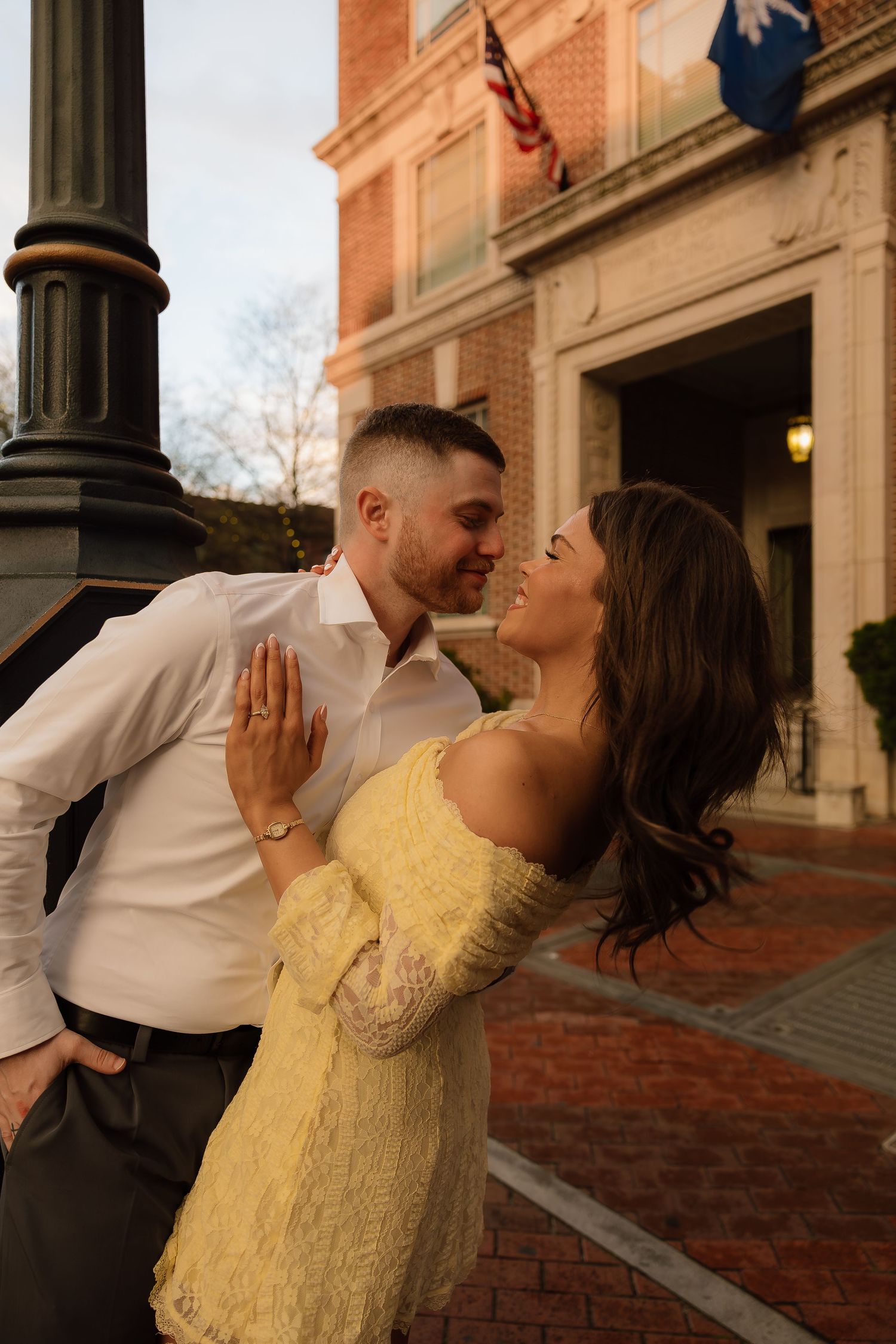 Couple sharing a romantic moment near a lamp post outside a brick building, woman in yellow lace dress smiling up at man.