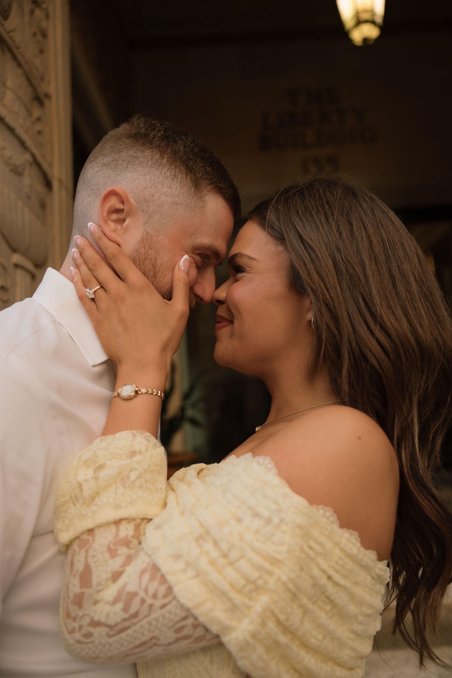 Romantic couple in close embrace, woman in off-shoulder lace dress with engagement ring, sharing an intimate moment.