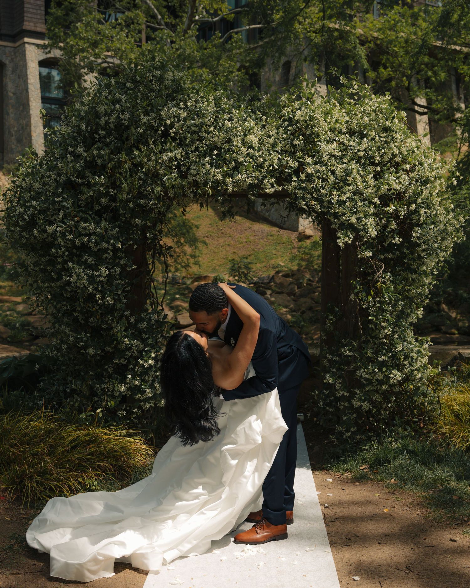 Bride and groom share a romantic dip kiss under a lush green floral arch on a sunlit garden pathway.