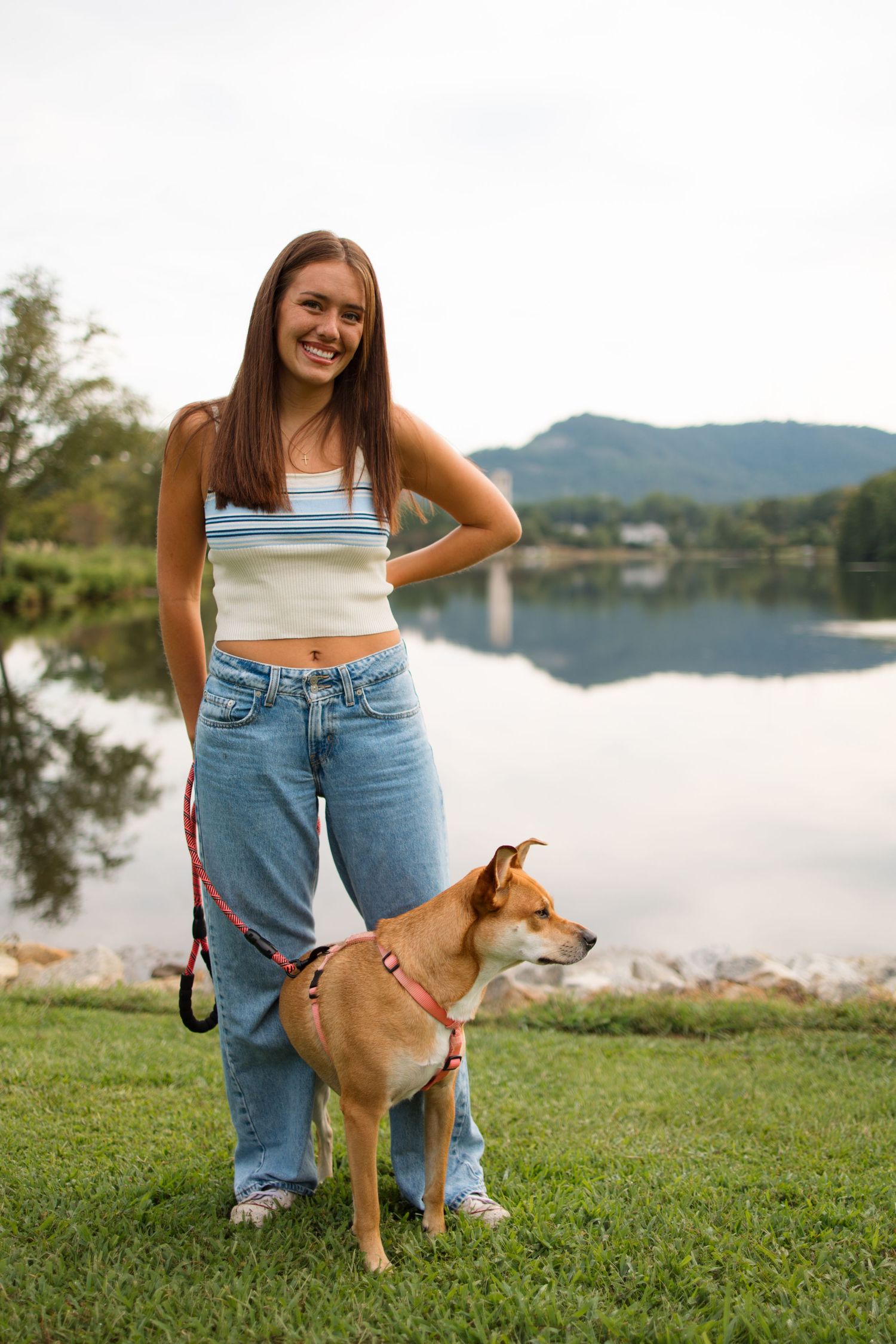 Person in casual denim poses with friendly tan dog on lakeside grass.