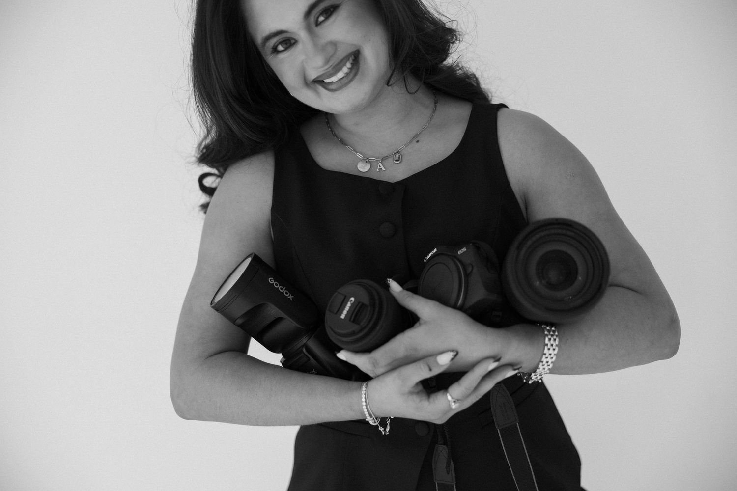 Black and white photo of a smiling woman holding multiple cameras, wearing a sleeveless top and bracelets.