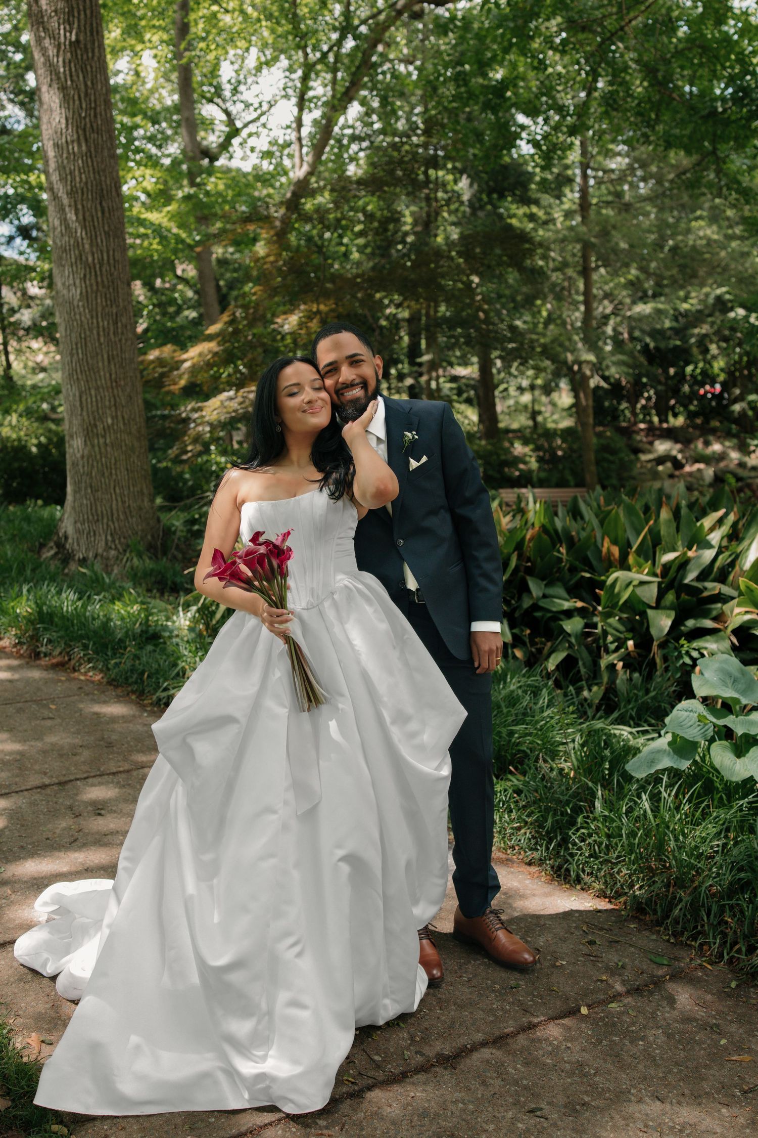 Bride in strapless white ballgown holding red bouquet poses with groom in navy suit in lush garden setting.