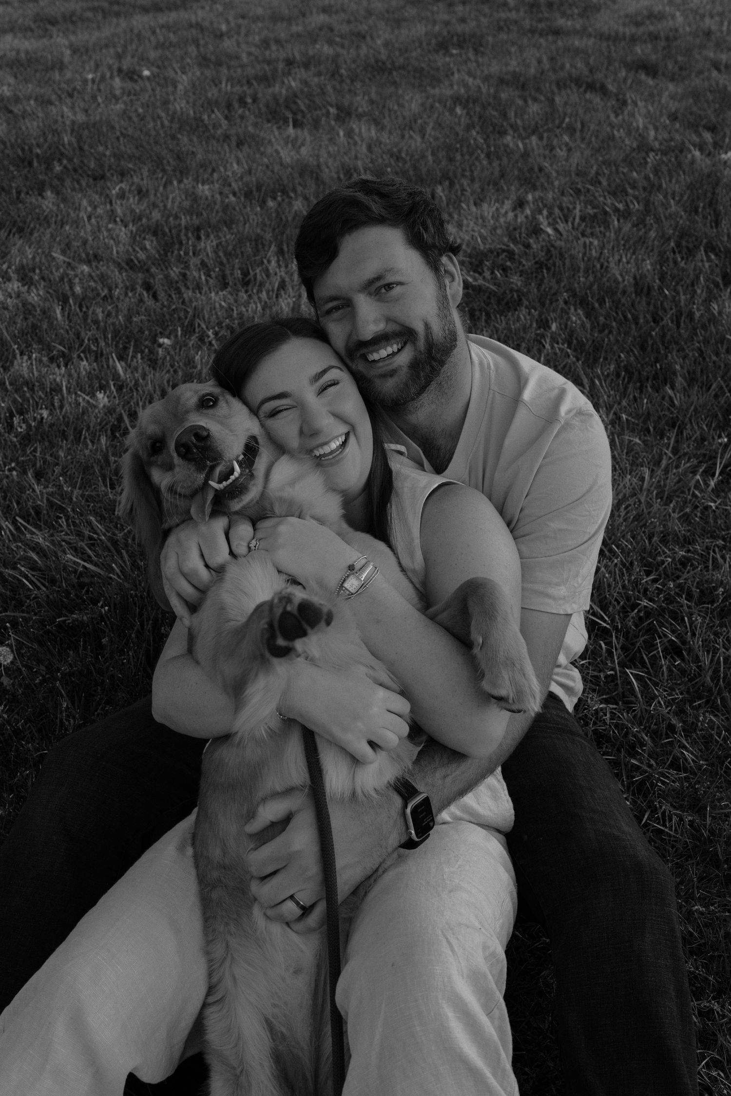Black and white photo of a smiling couple sitting on grass, joyfully hugging their happy golden retriever dog.