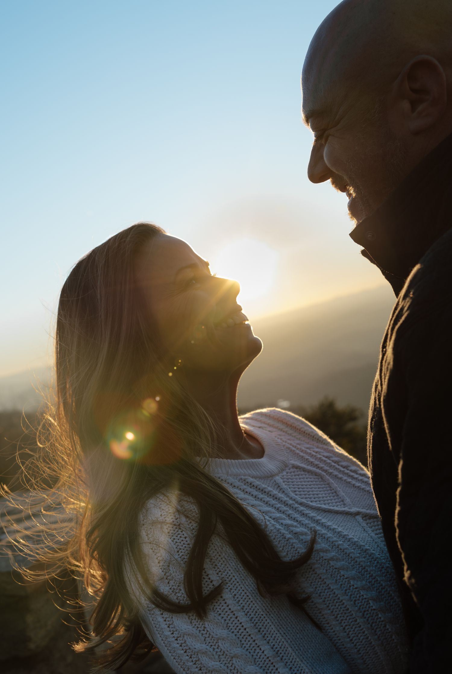A couple embraces in silhouette against a golden sunset with lens flare creating a romantic backlit glow.