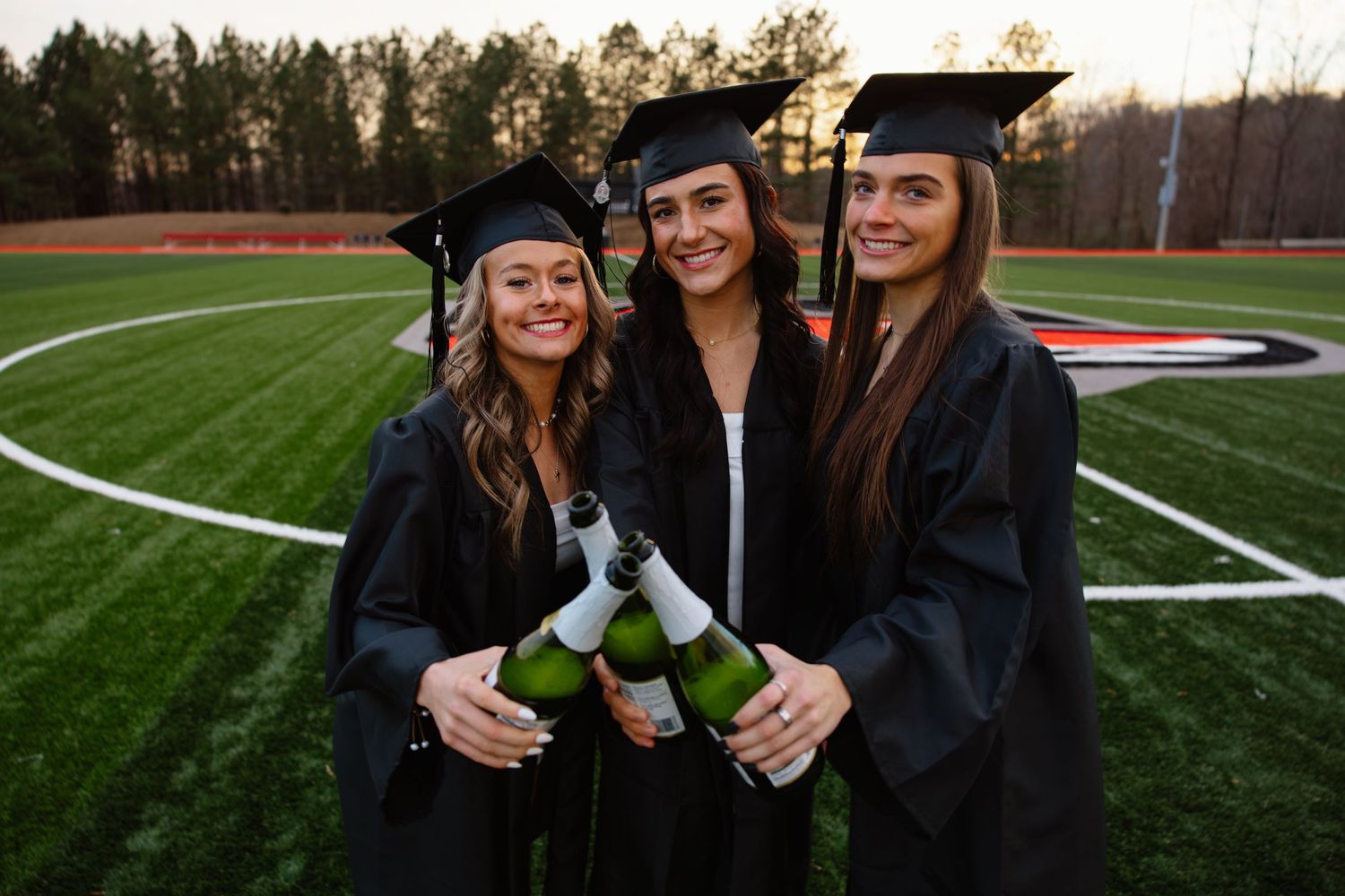 Three graduates in caps and gowns celebrate with diplomas on a sports field.