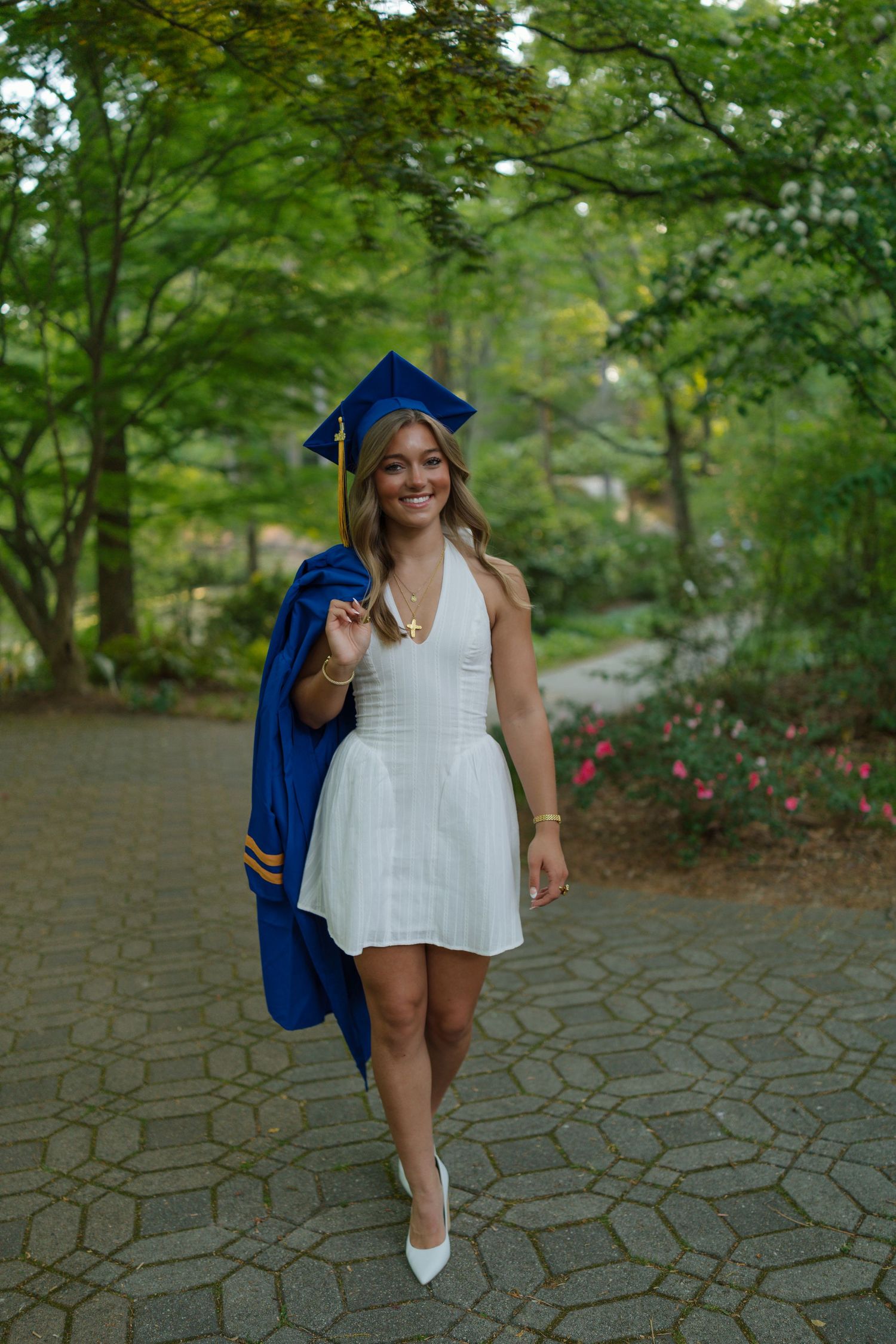 Smiling graduate in white dress and blue cap and gown walking along a tree-lined pathway.