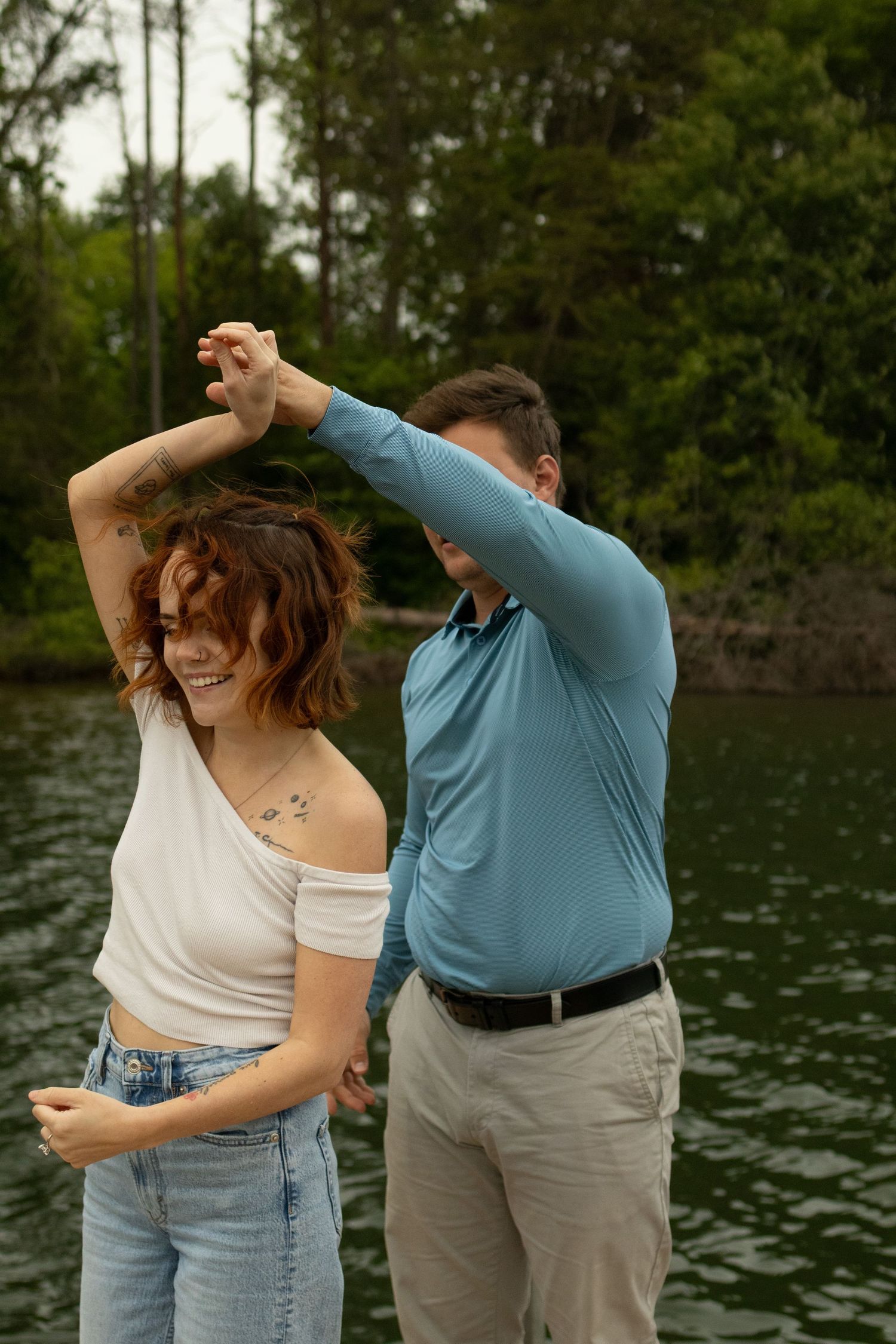 A couple shares a joyful moment dancing together by a serene lake surrounded by trees.