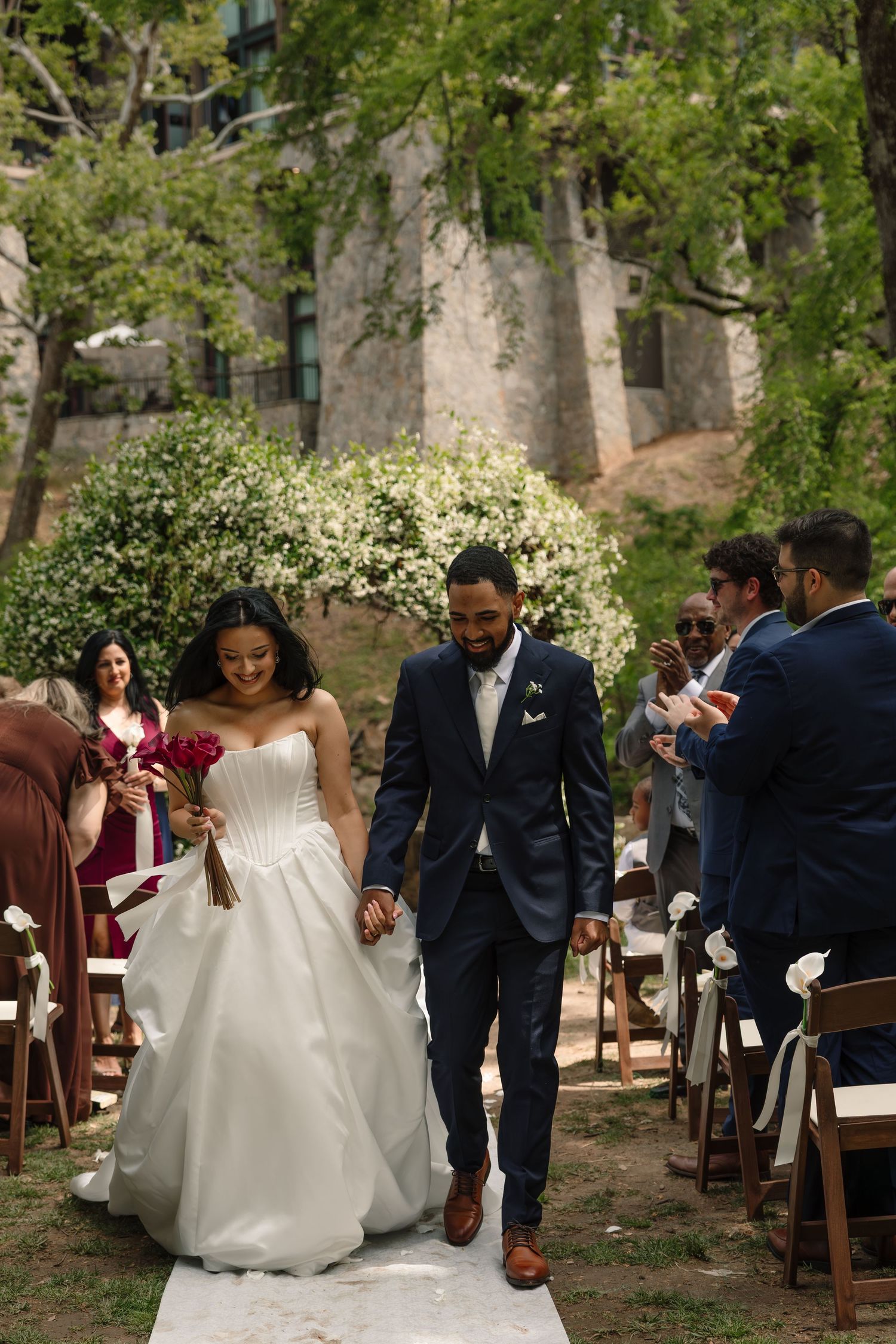 Bride and groom walk down outdoor aisle after wedding ceremony, guests applauding, lush greenery and stone building backdrop.