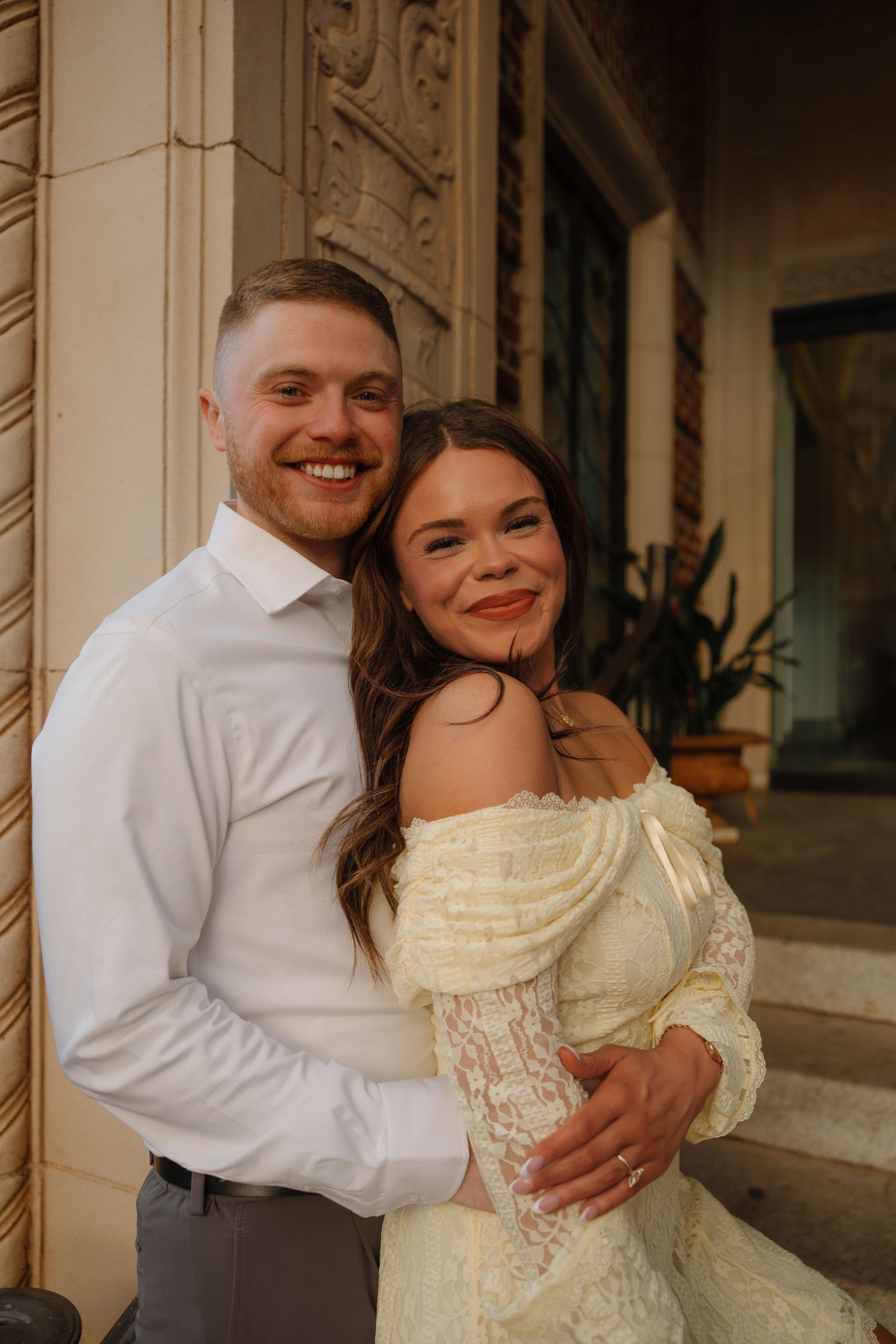 A smiling couple poses outdoors, the man in a white shirt embracing a woman in an off-shoulder lace dress.