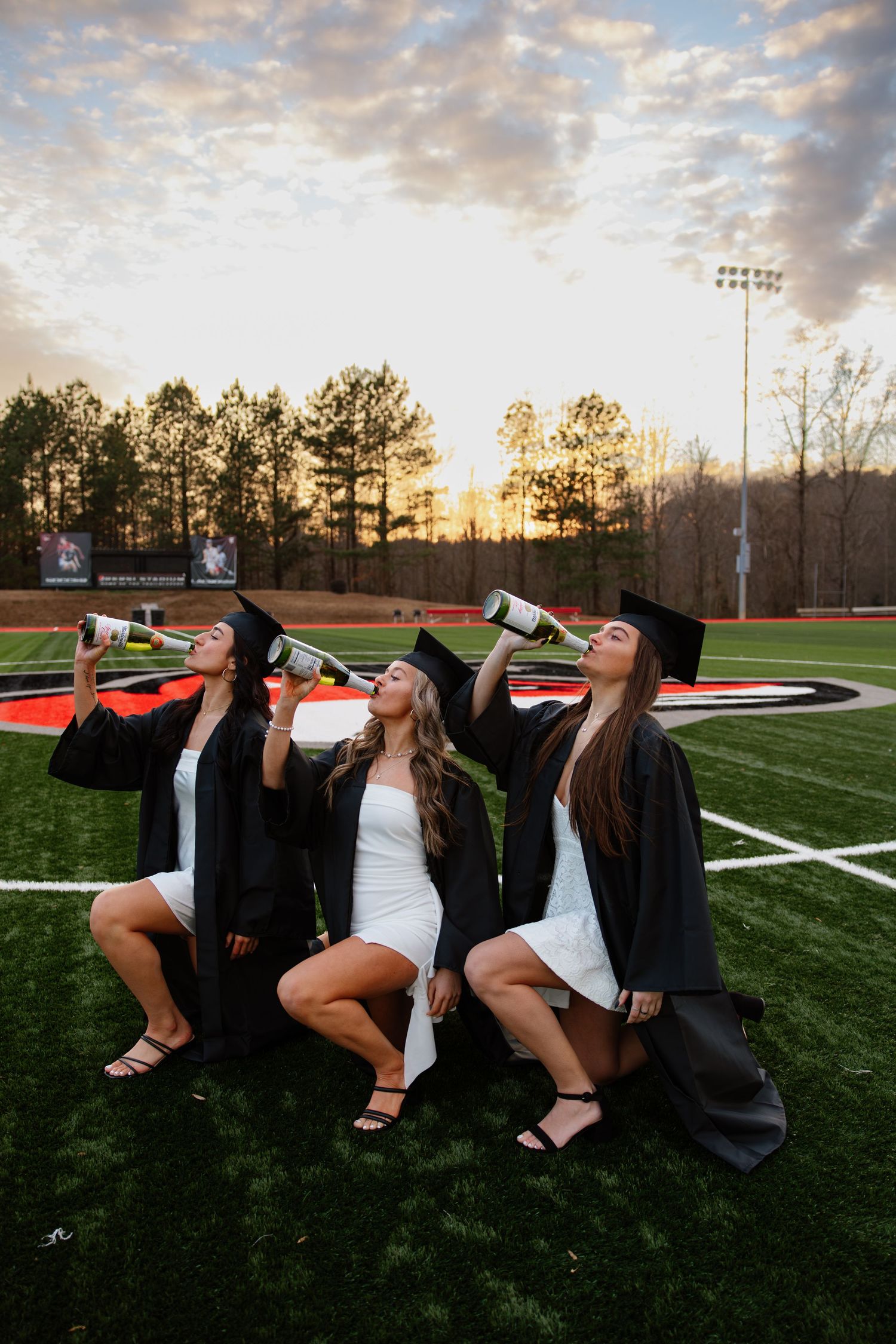 Friends in graduation attire lying on grass field drinking from bottles during sunset celebration.