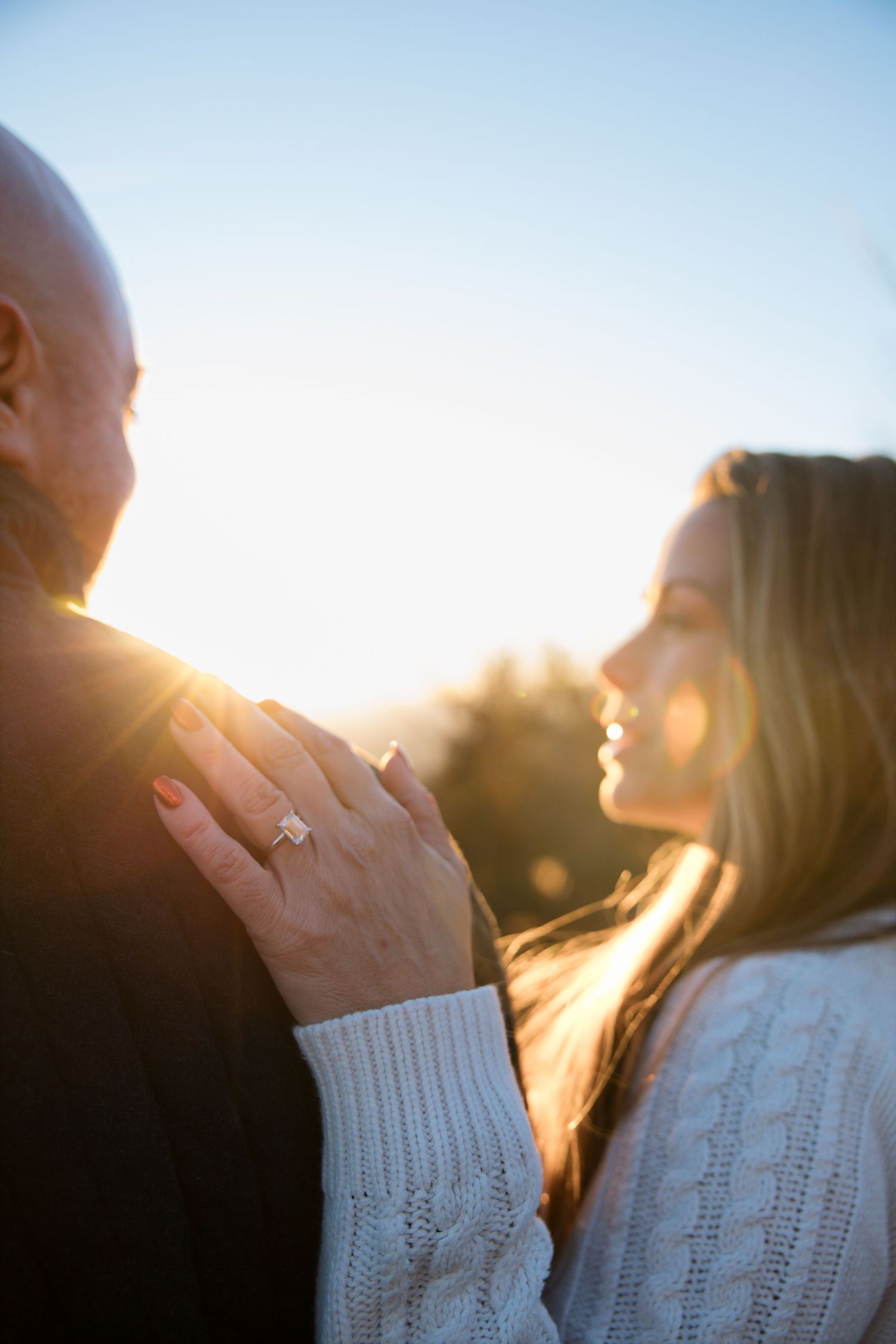 A couple embraces during golden hour sunlight, with the woman wearing a white knit sweater and holding hands tenderly.
