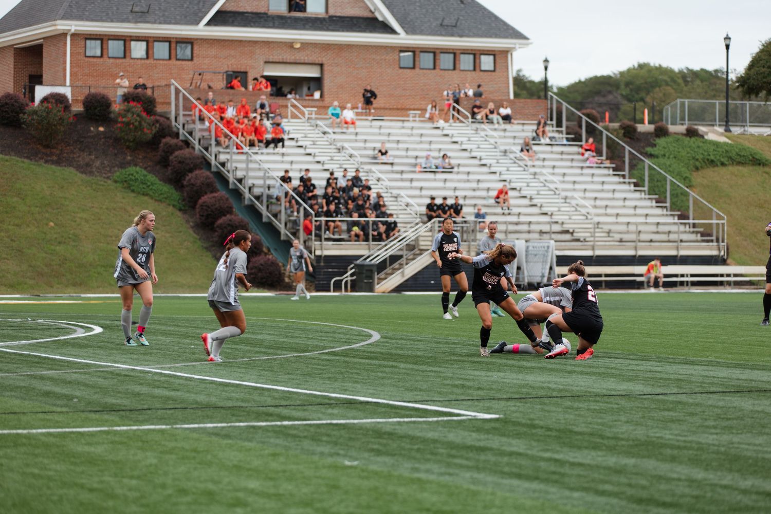 Soccer match action on green turf field with spectators watching from bleachers.