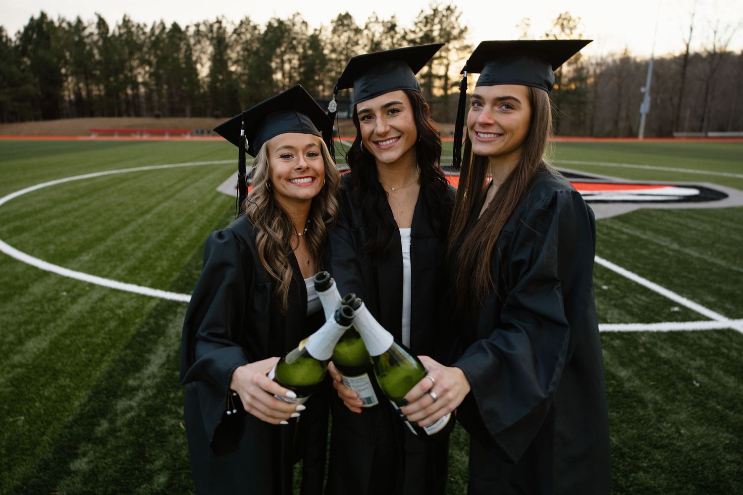 Three graduates in black caps and gowns celebrating with champagne bottles on sports field.