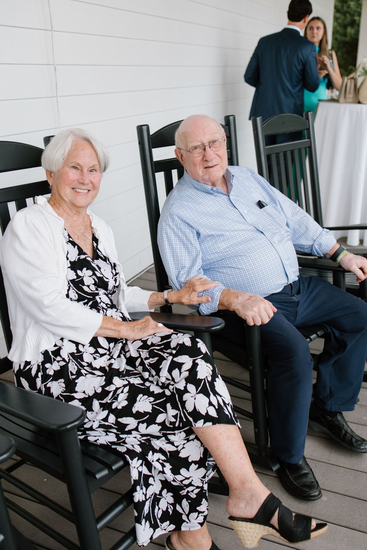 Elderly couple relaxing together on a porch in black and white floral dress and casual attire.