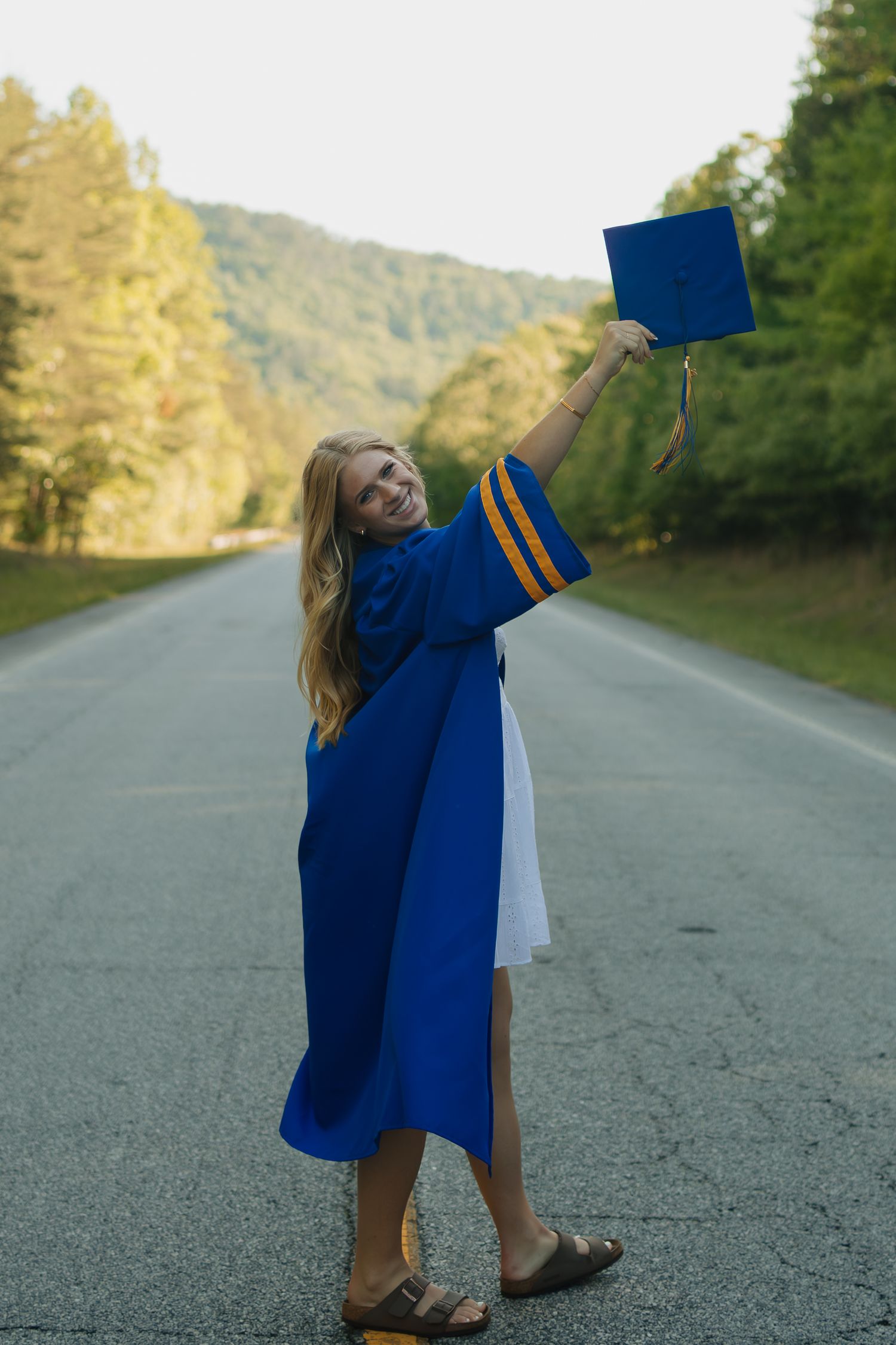 Young woman in blue graduation cap and gown celebrating on a tree-lined road, holding her cap high in the air.