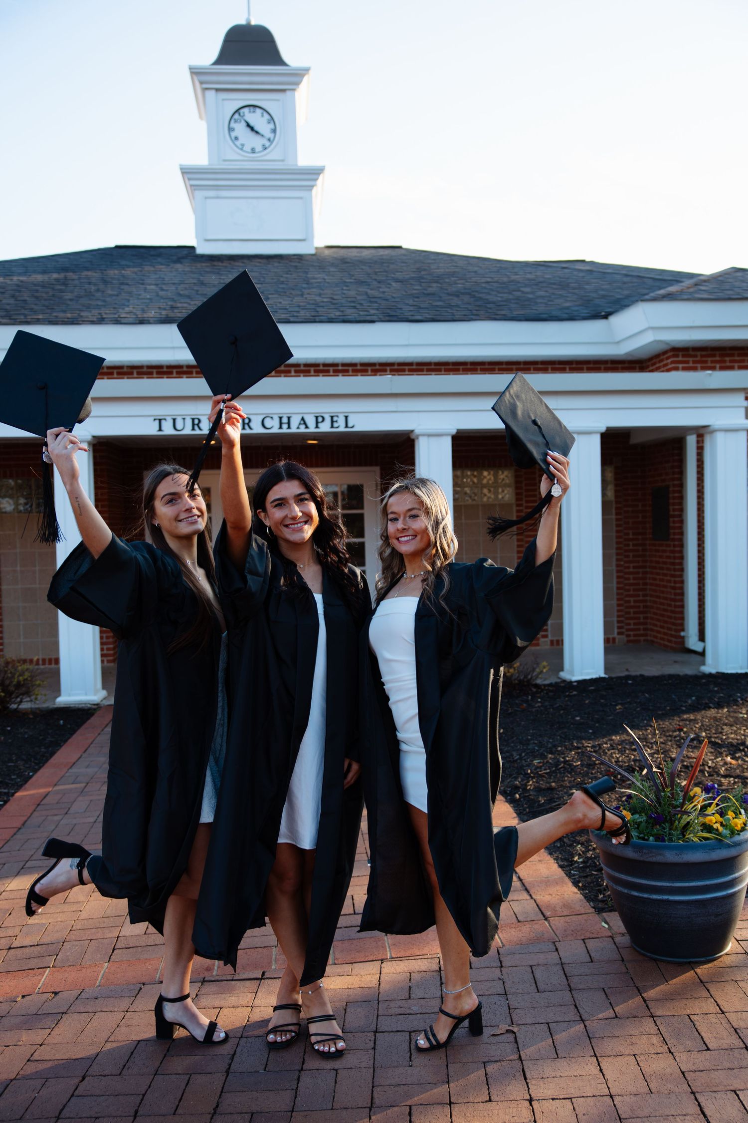 Three graduates in black robes celebrate by tossing caps in front of a chapel building.