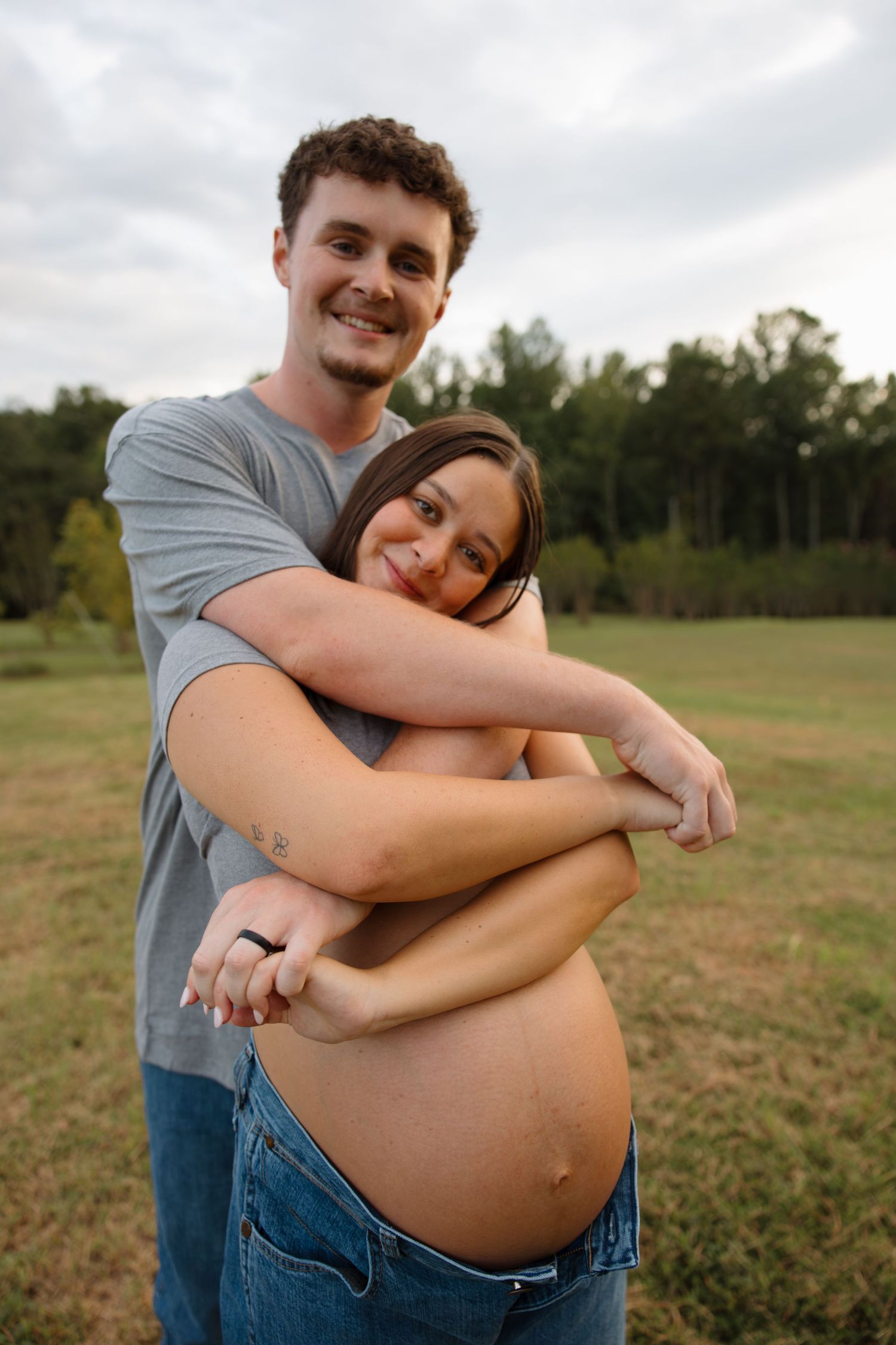 Couple sharing playful embrace in sunny field during maternity shoot.