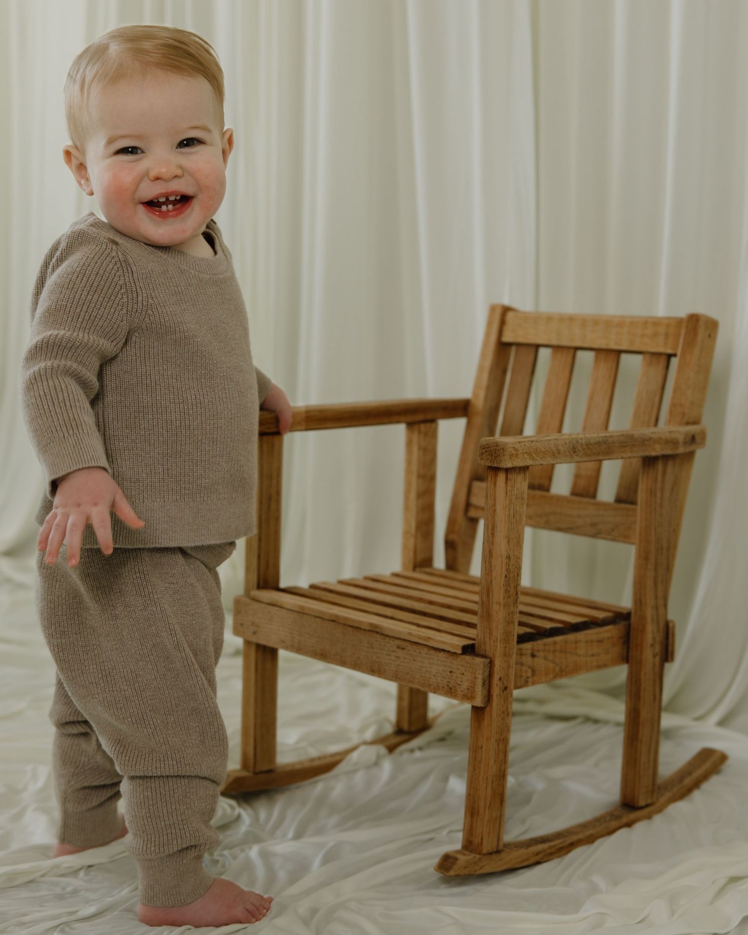 Happy toddler in beige outfit standing next to wooden rocking chair against white curtains.