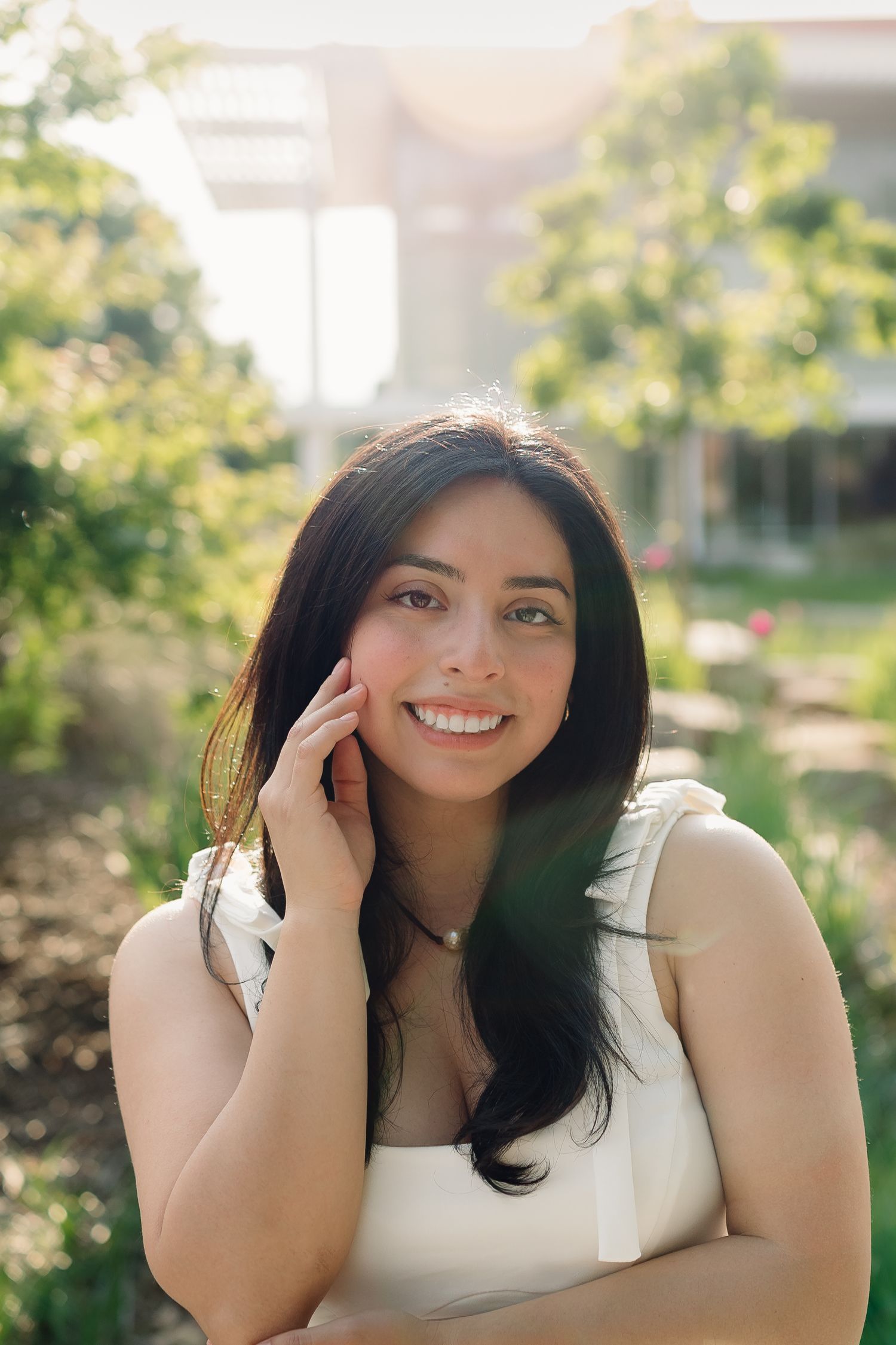 Smiling young woman with dark hair in a white top posing outdoors in a sunny green garden setting.