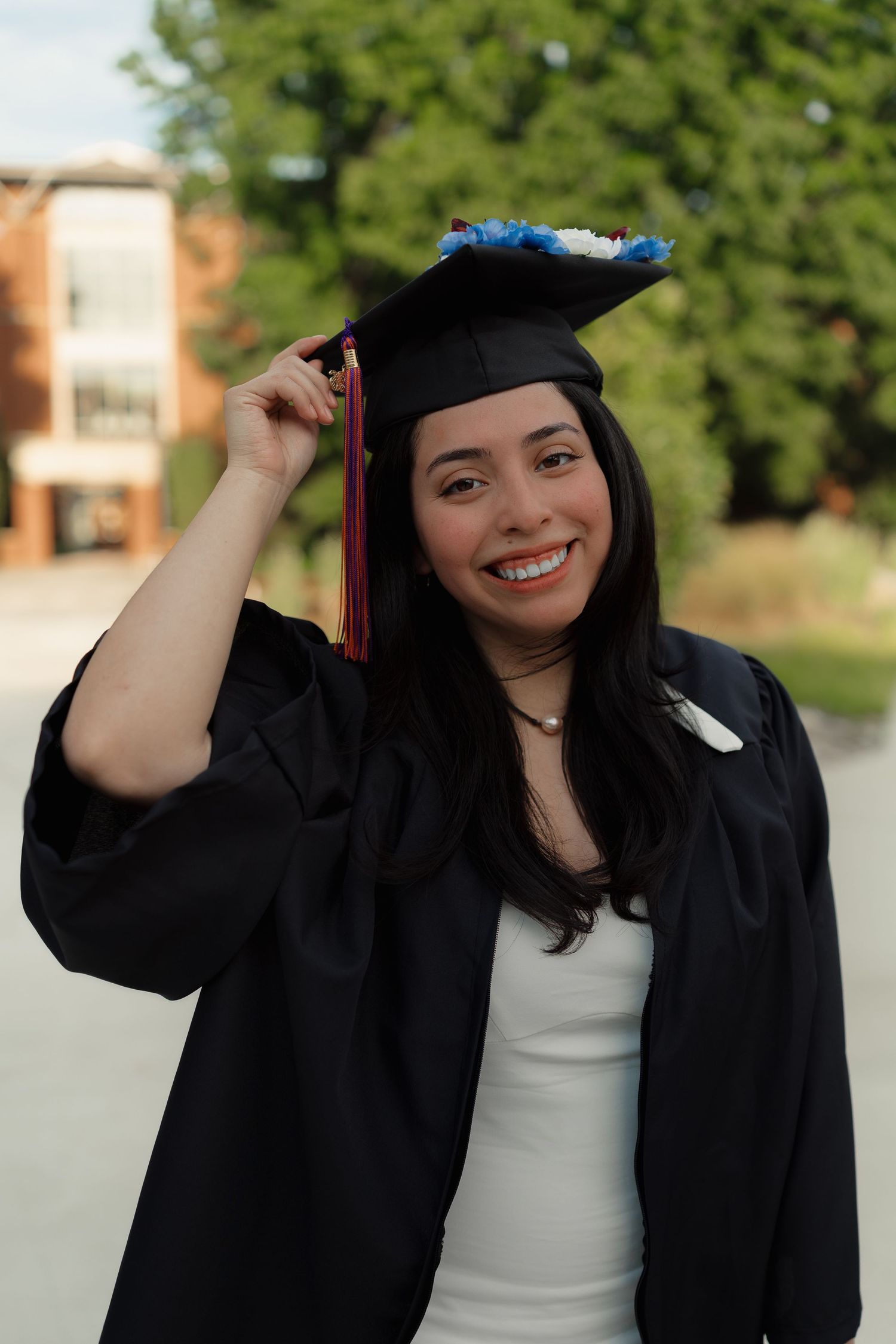 Smiling young woman in black graduation gown and cap holding tassel outdoors on college campus.