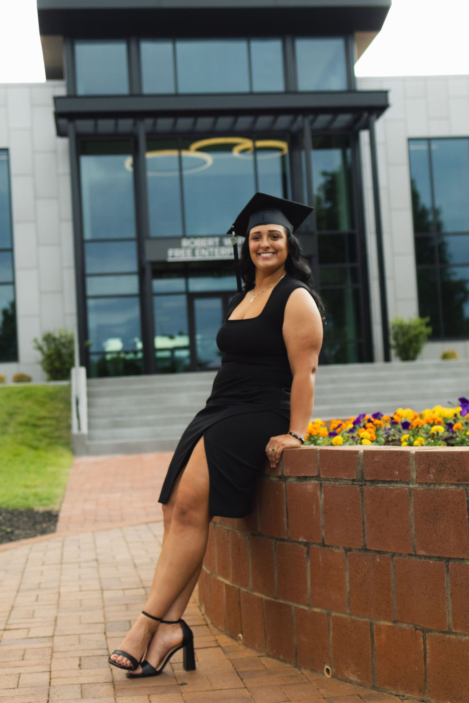 Graduate in black dress sitting on brick wall near modern building with flowers.