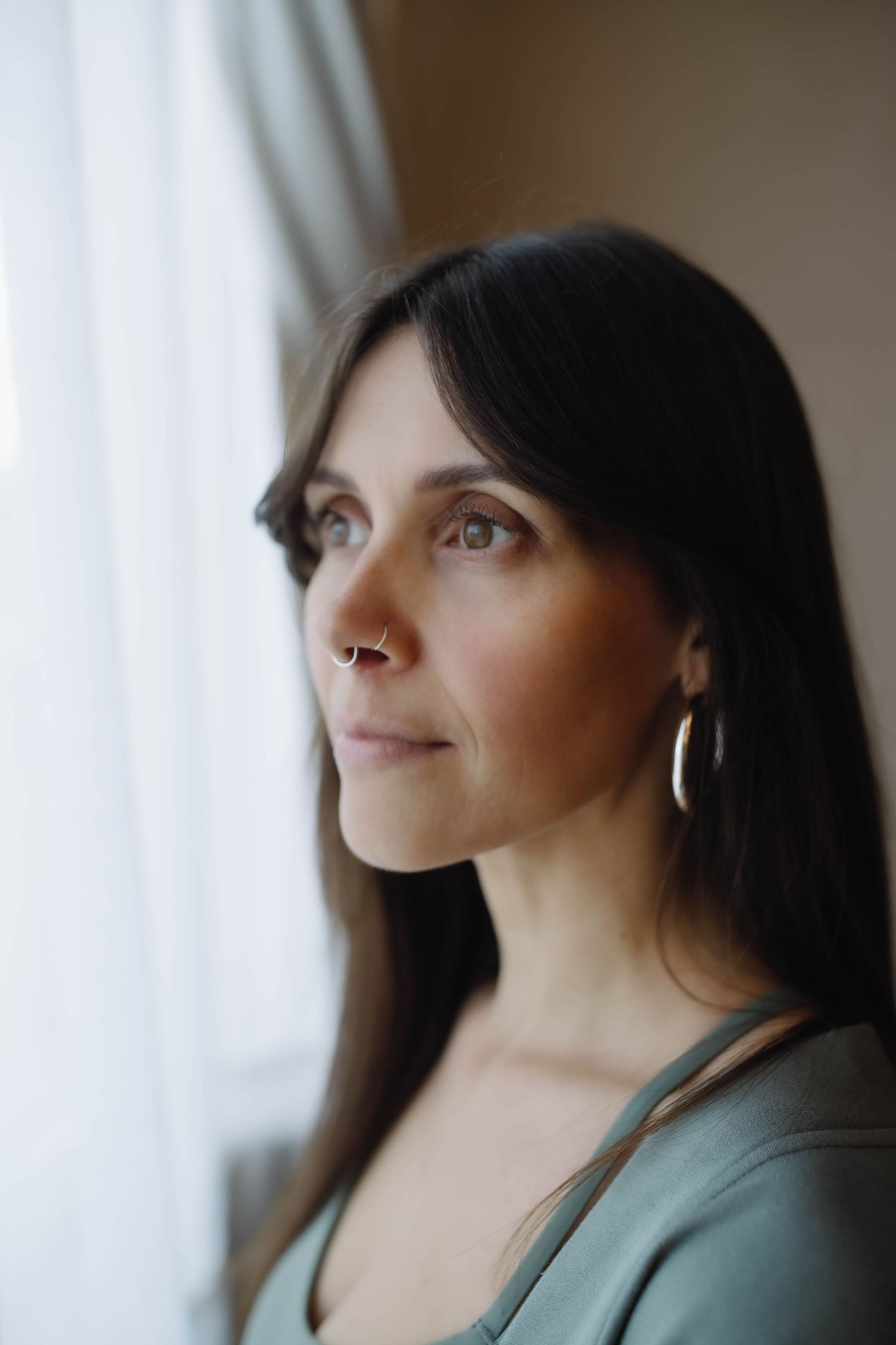 A thoughtful brunette woman with hoop earrings and a nose ring gazes upward near a bright window in soft light.