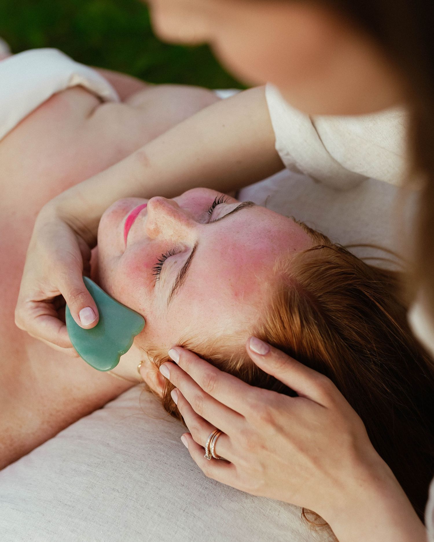 Esthetician performing a gua sha facial massage with a jade stone tool on a relaxed woman lying outdoors.